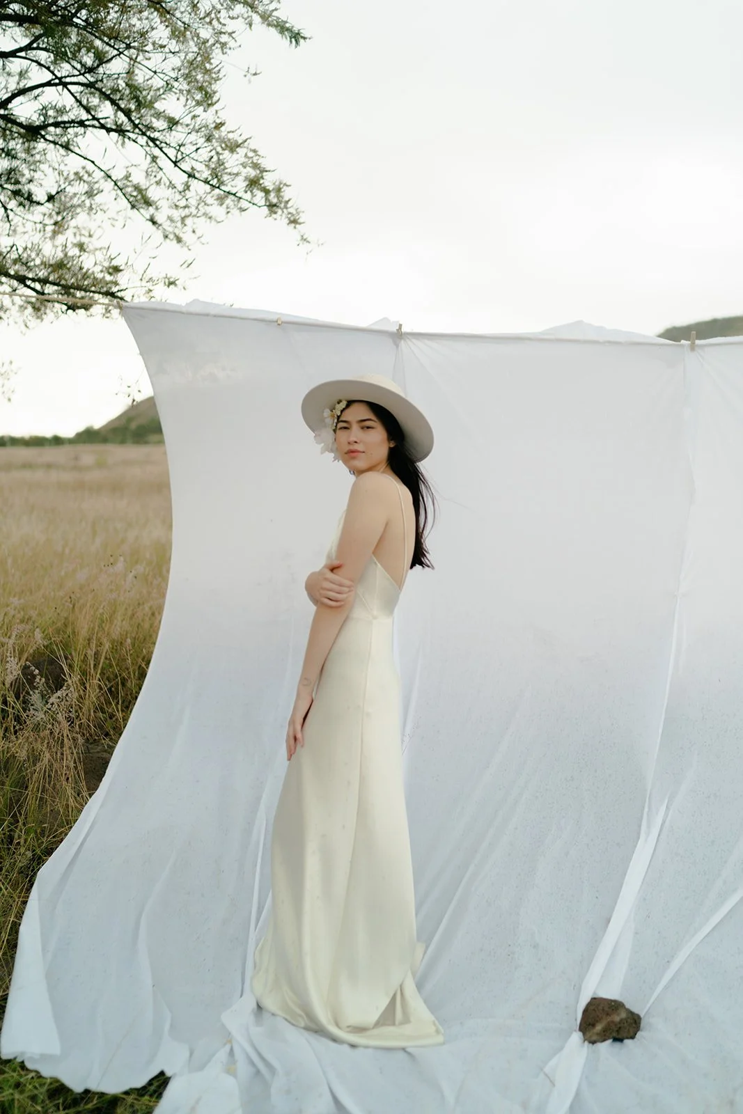 Woman in white dress and hat standing outdoors against a white fabric backdrop with grassy field background.