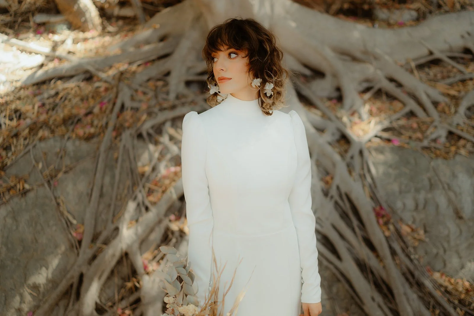Person in white dress standing in front of large tree roots with dry leaves and twigs on the ground, looking to the side, wearing floral earrings.