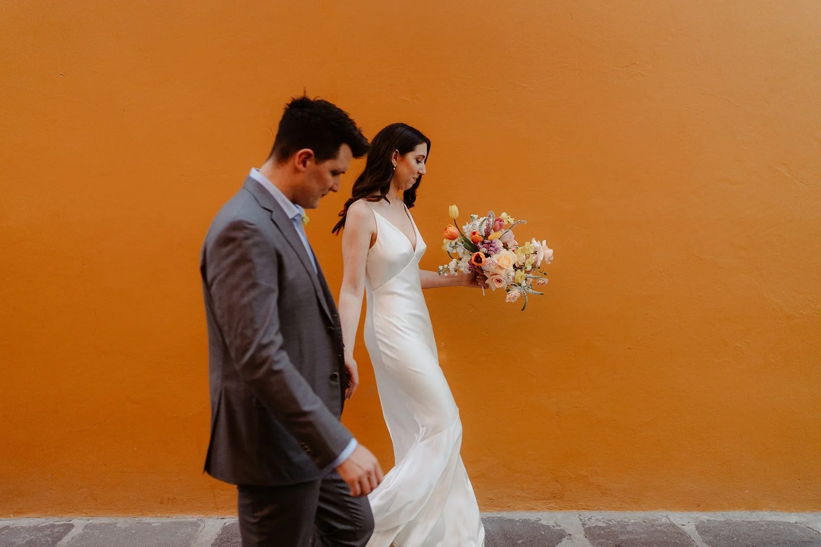 A bride in a white dress holding a colorful bouquet walks alongside a man in a gray suit against an orange wall backdrop.