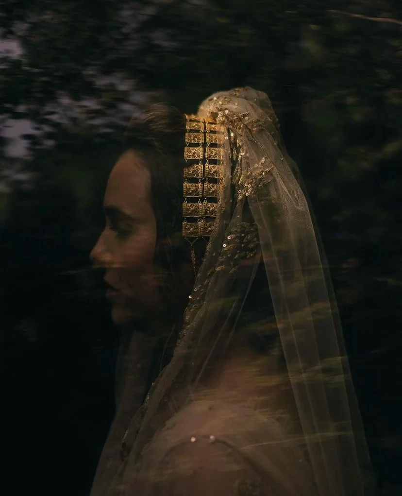 Profile of a woman wearing a decorative gold headpiece and sheer veil, surrounded by dark, blurred foliage.