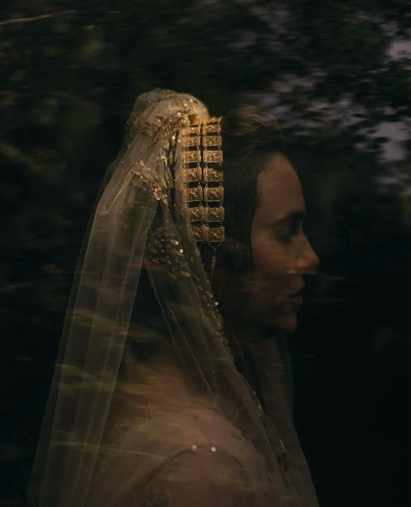 A person wearing a decorative headpiece and veil in dim lighting, surrounded by a dark background with blurred foliage.