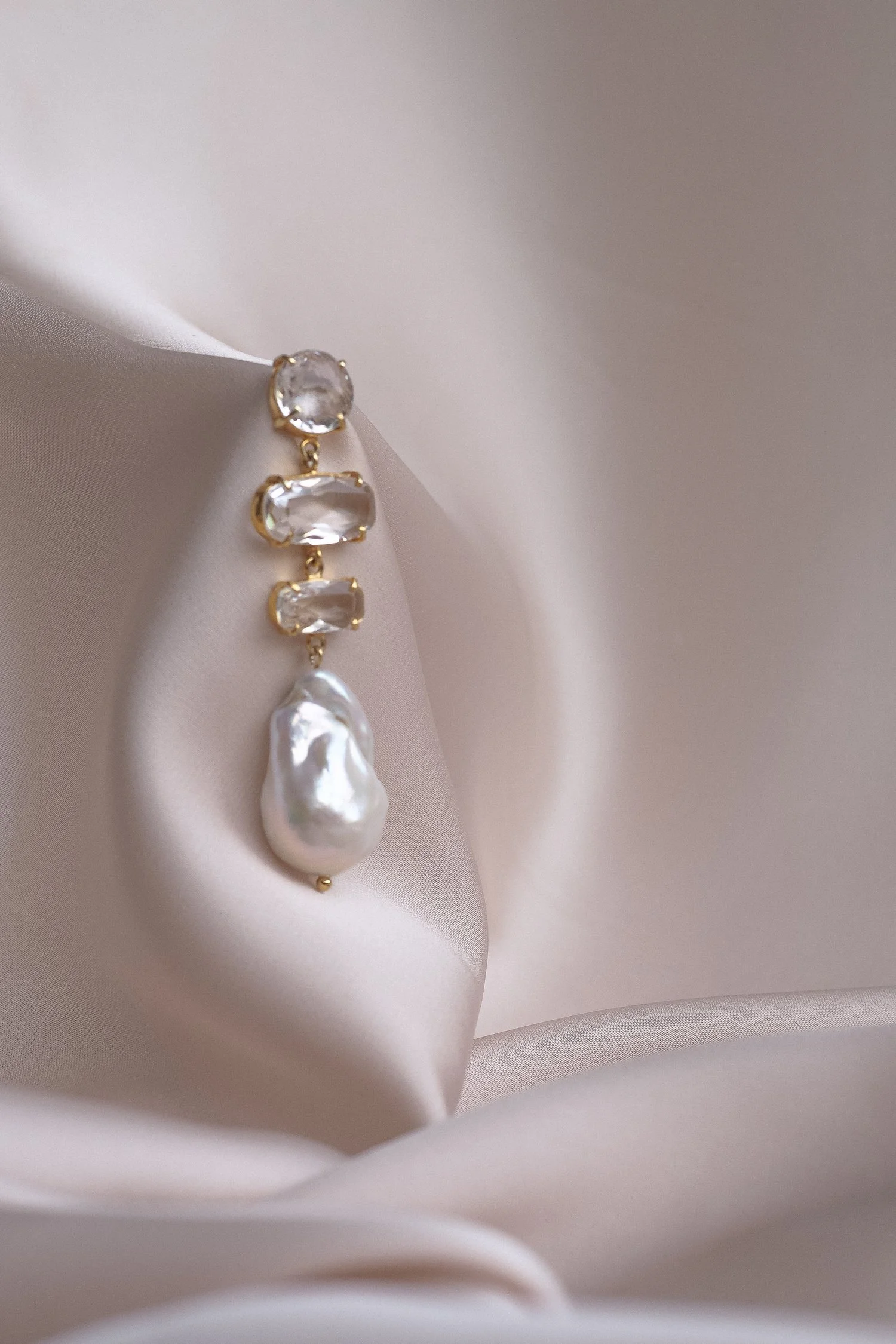 Elegant gold earring with clear gemstones and a baroque pearl on a soft beige fabric background.