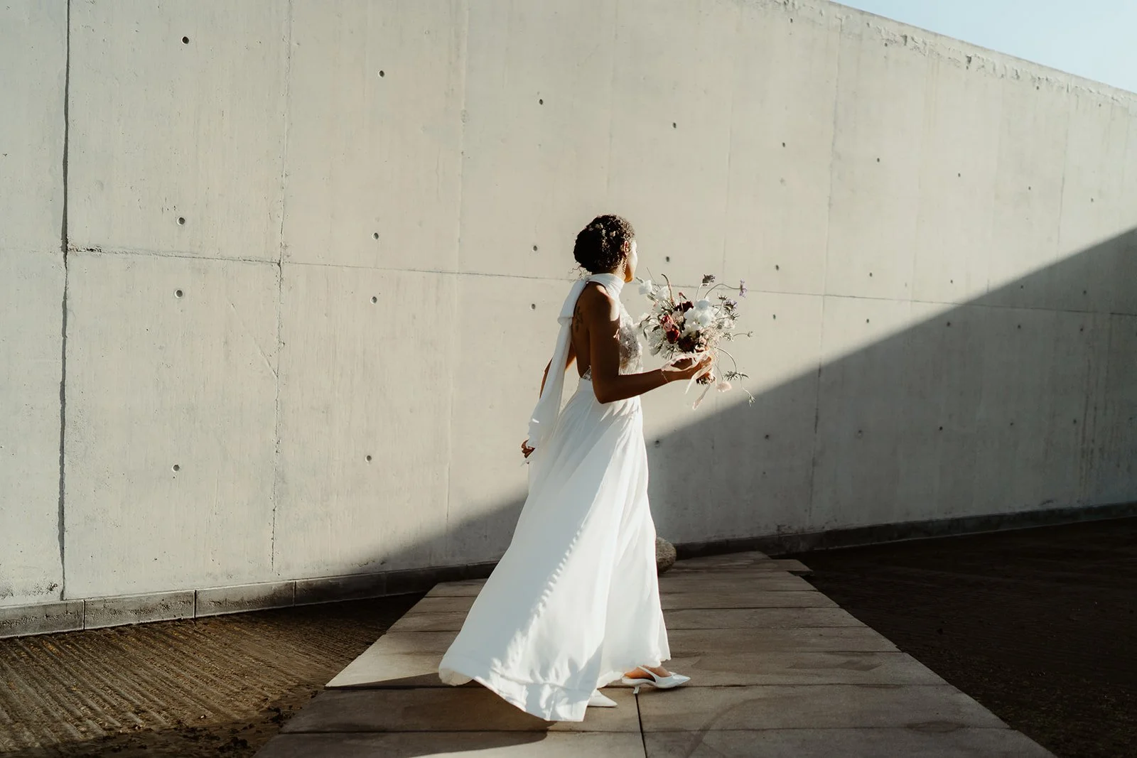 Bride in white dress holding bouquet walking near concrete wall