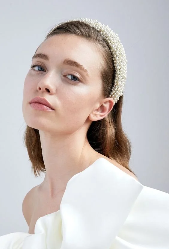 Woman wearing a pearl headband and off-shoulder white dress against a light backdrop.