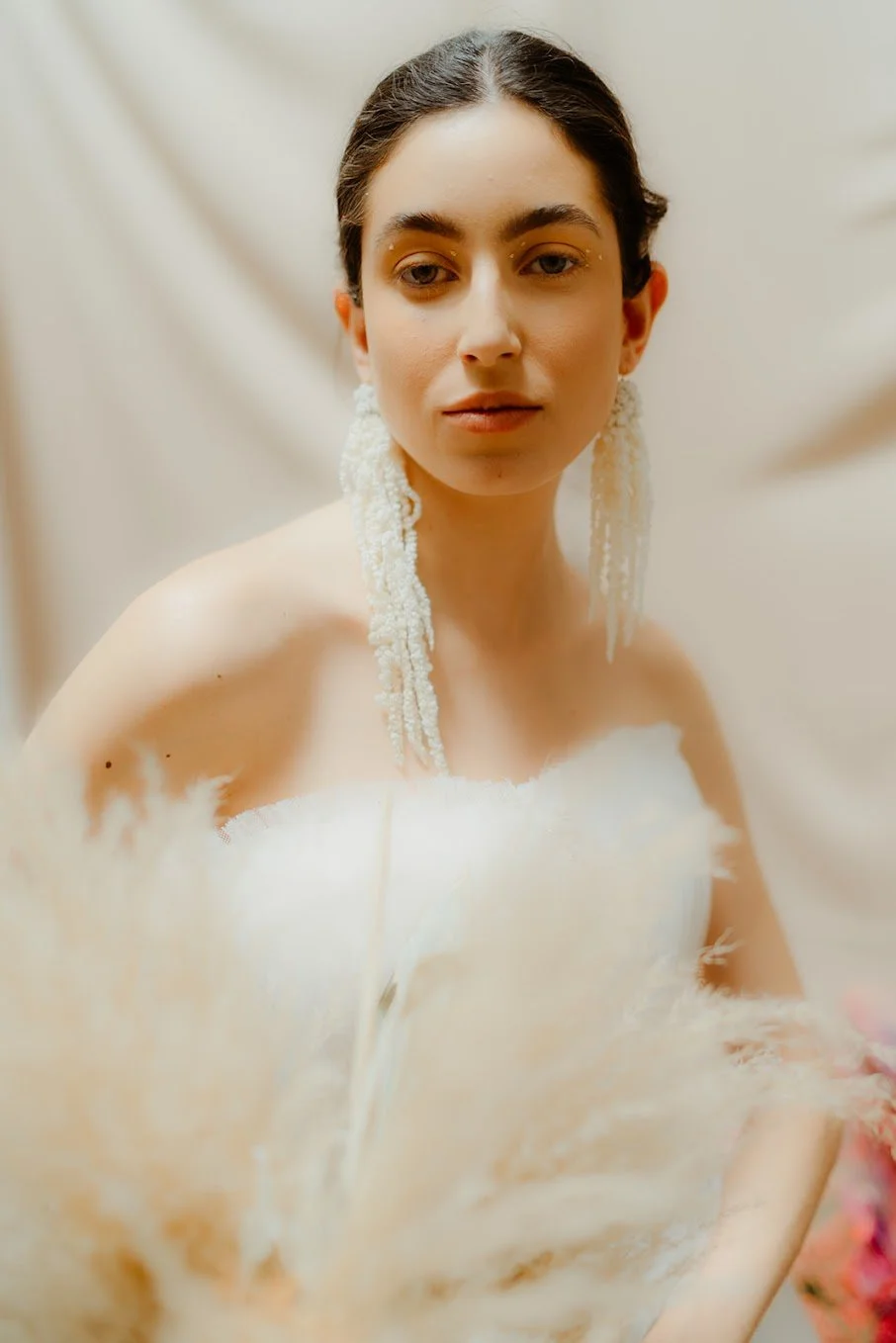 Woman wearing a strapless white dress, large beaded earrings, holding pampas grass.