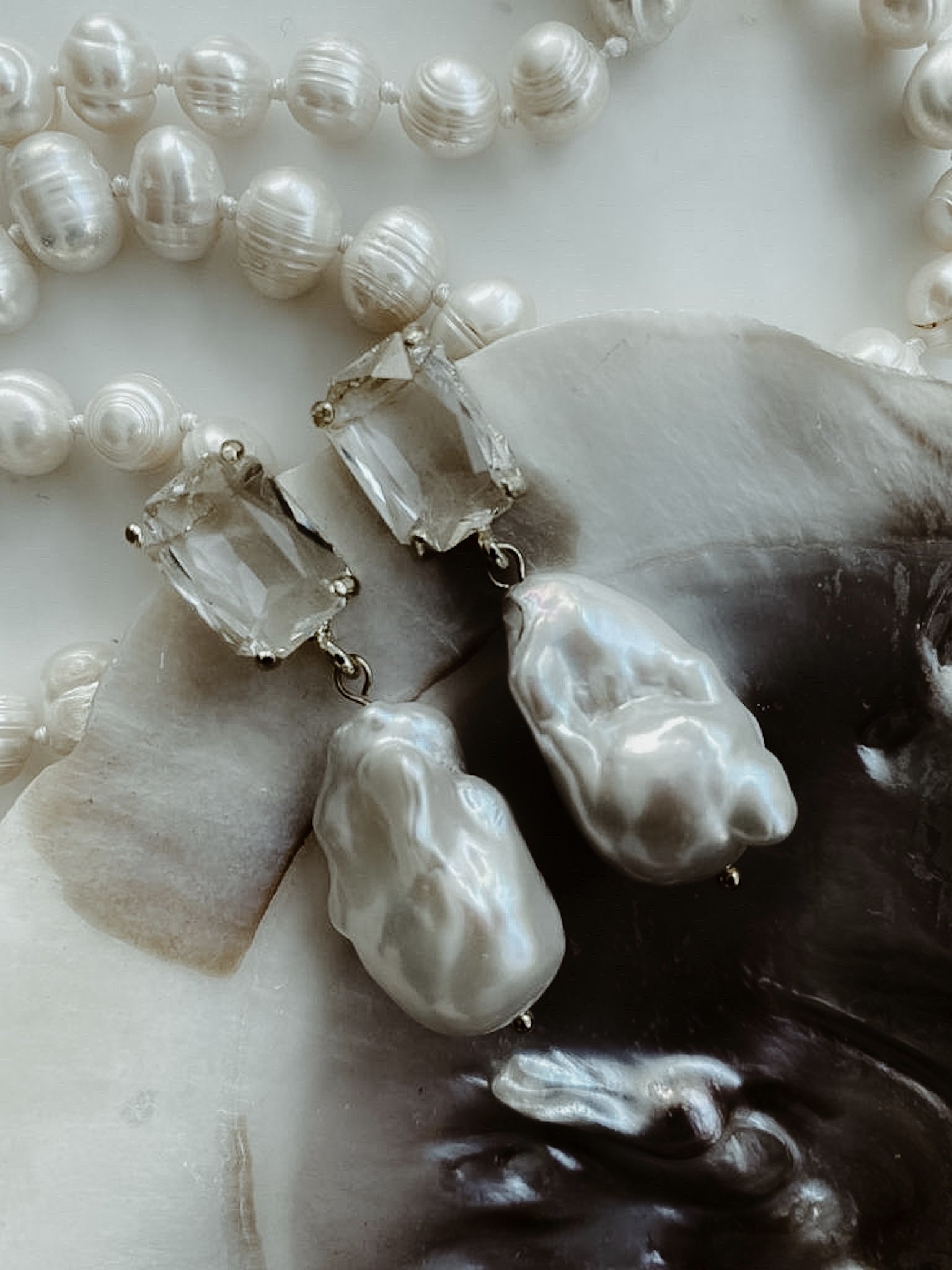 Baroque pearl earrings with crystal studs on a background of pearl necklaces and an oyster shell.