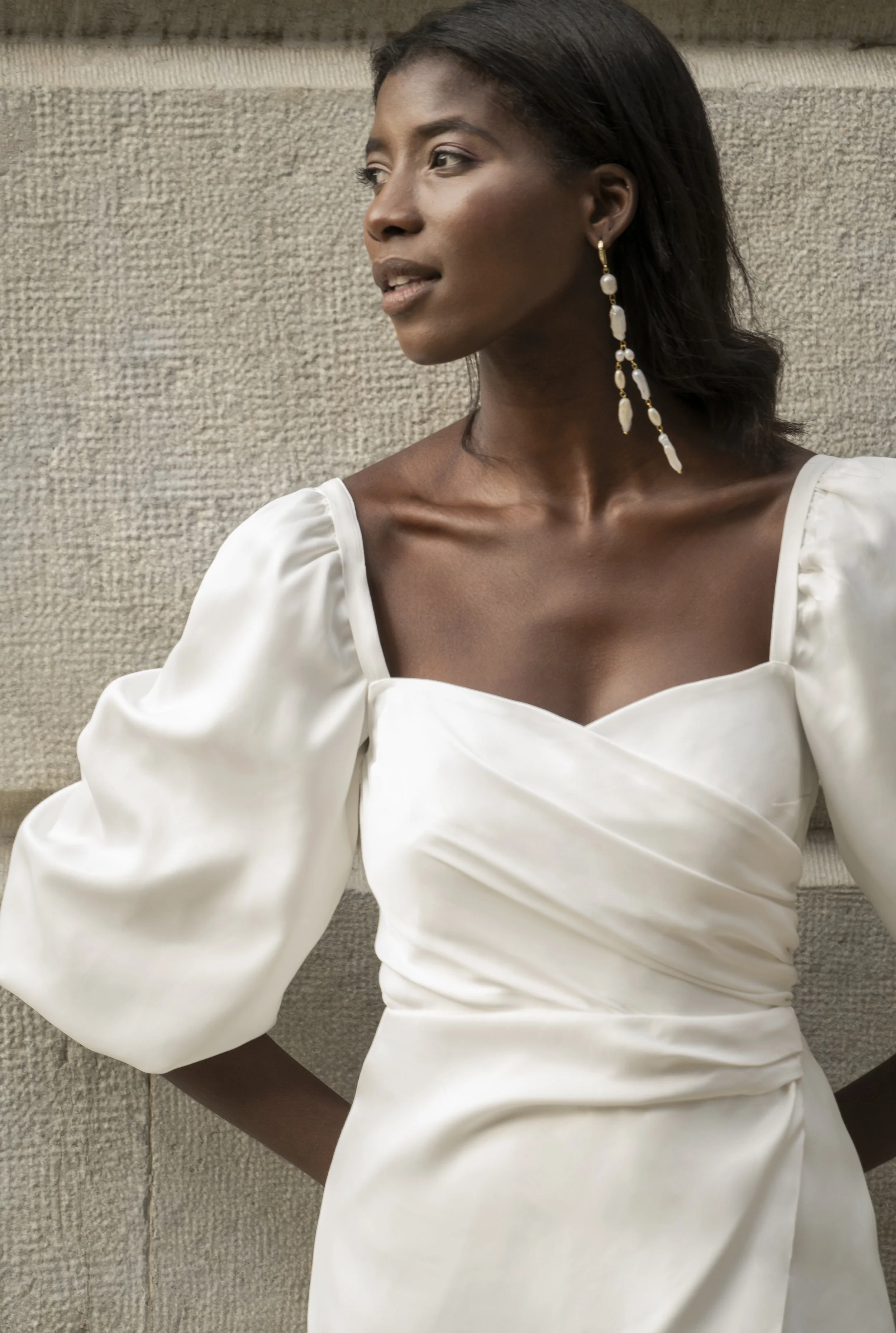 Person wearing an elegant white dress with puffed sleeves and long hanging earrings, standing against a textured wall.