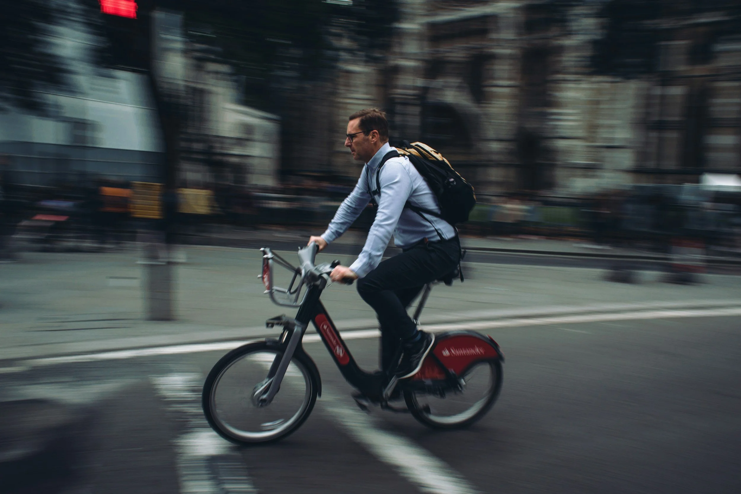 Man wearing glasses, light blue shirt, dark pants, riding a red bicycle on a city street during daytime, with buildings and blurred pedestrians in the background