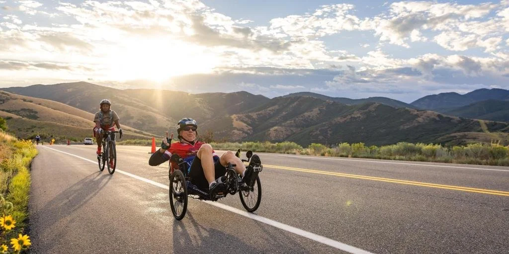 A person riding a handcycle on a scenic rural road with mountains in the background, during sunset. Another cyclist follows, and there are traffic cones on the road.