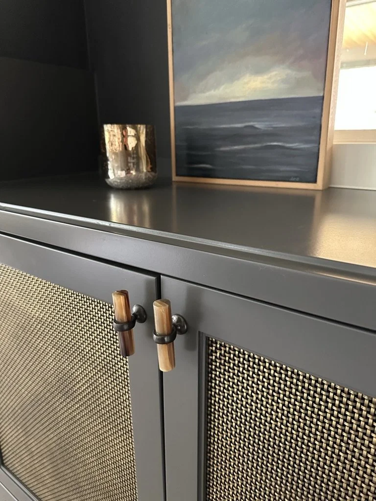 Gray cabinet with woven panels, wooden handles, and a small framed seascape painting on top, alongside a reflective candle holder.