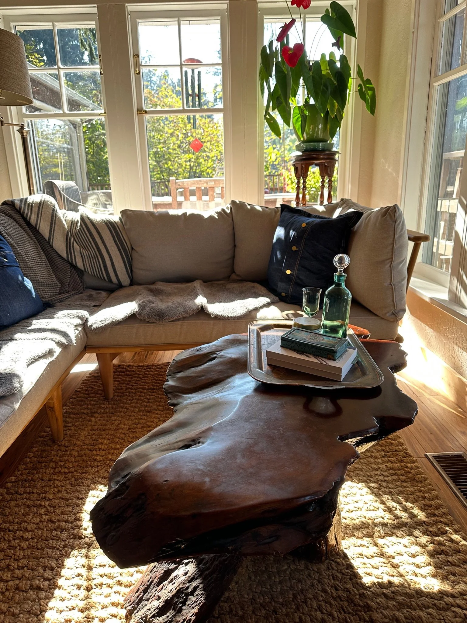 Cozy living room corner with a rustic wooden coffee table, beige sectional sofa with pillows, and large windows. A potted plant sits on a wooden stand. Sunlight illuminates the space.