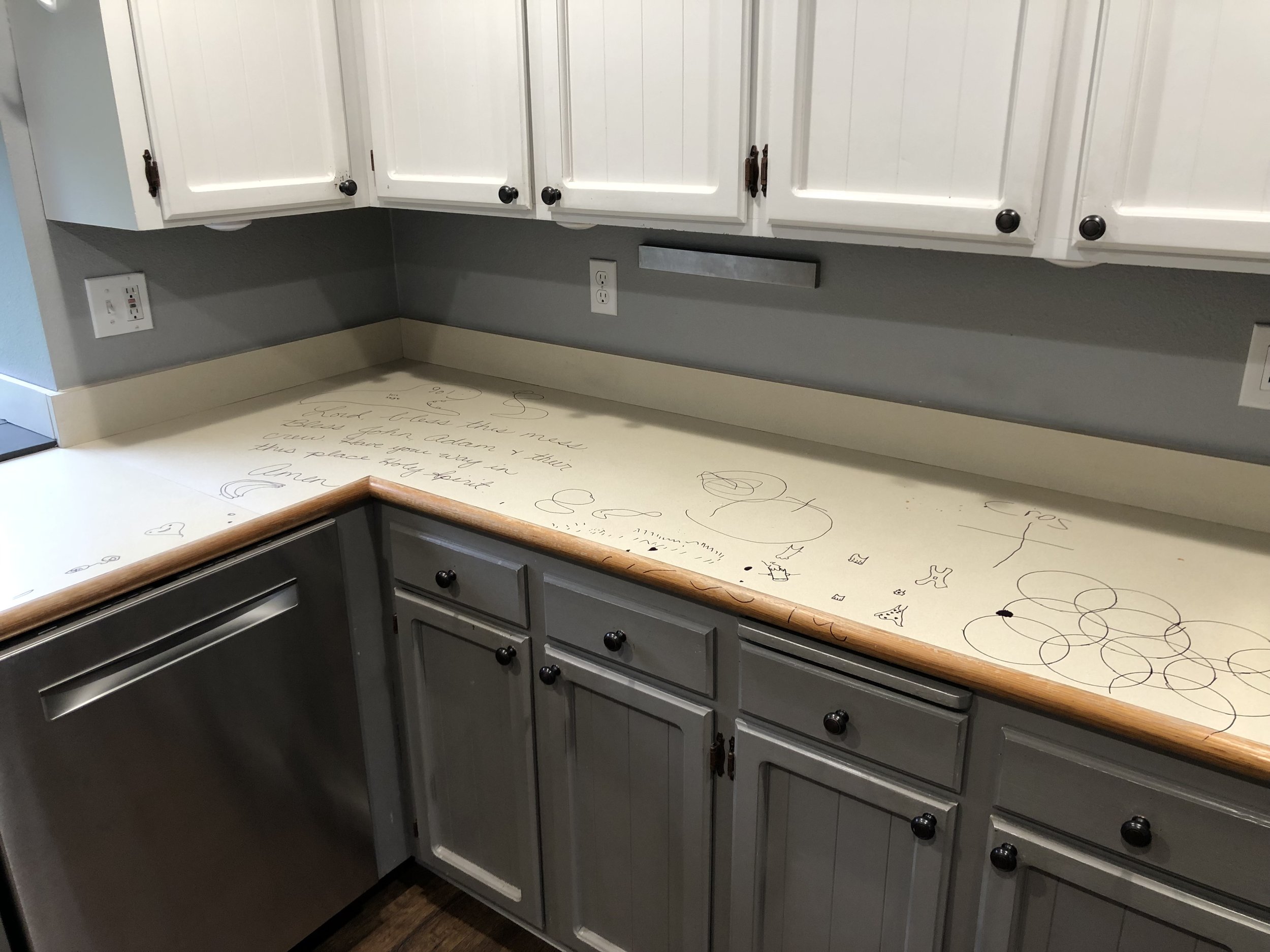 Kitchen countertop with hand-drawn doodles and messages, surrounded by gray cabinets and a stainless steel dishwasher.
