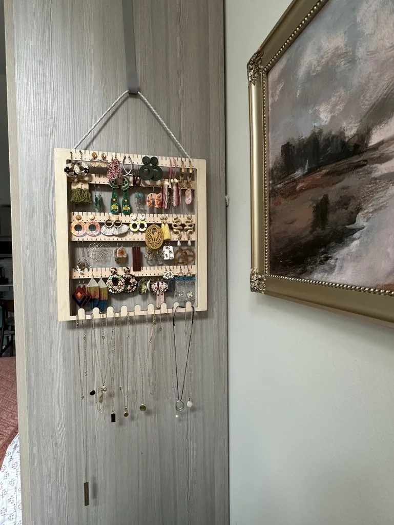 Wall-mounted jewelry holder with earrings and necklaces displayed next to a framed painting.