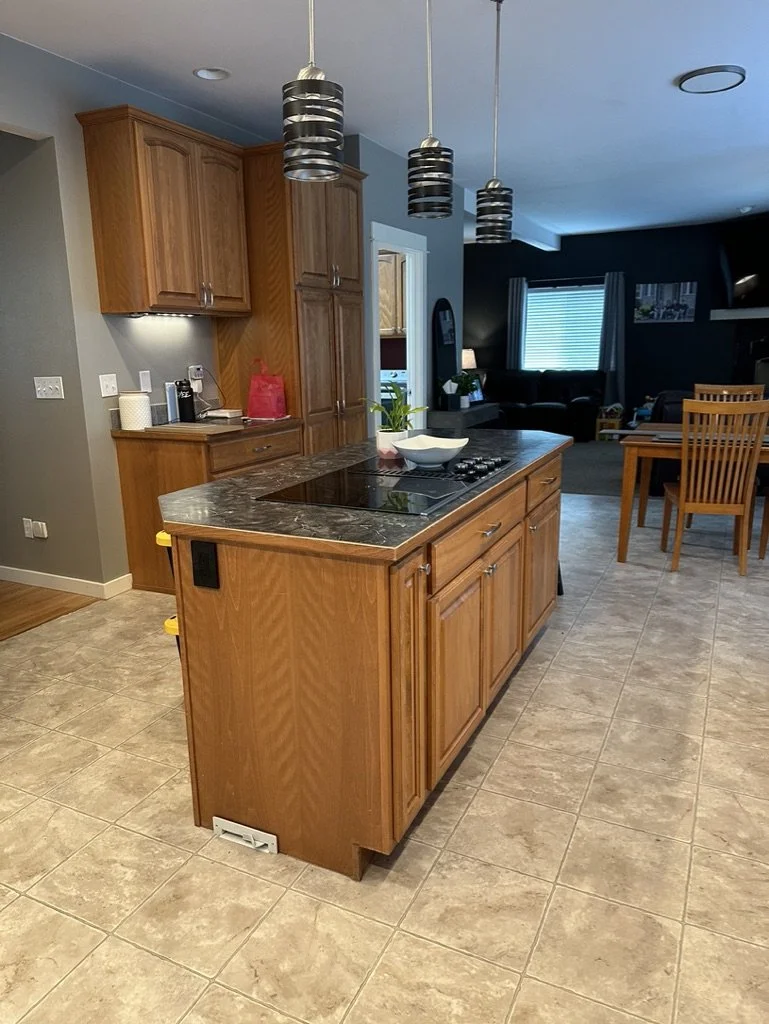Modern kitchen with wooden cabinets, a marble countertop island, pendant lights, and tiled flooring. In the background, there is a dining area with wooden chairs and a visible living room area with a television and couches.