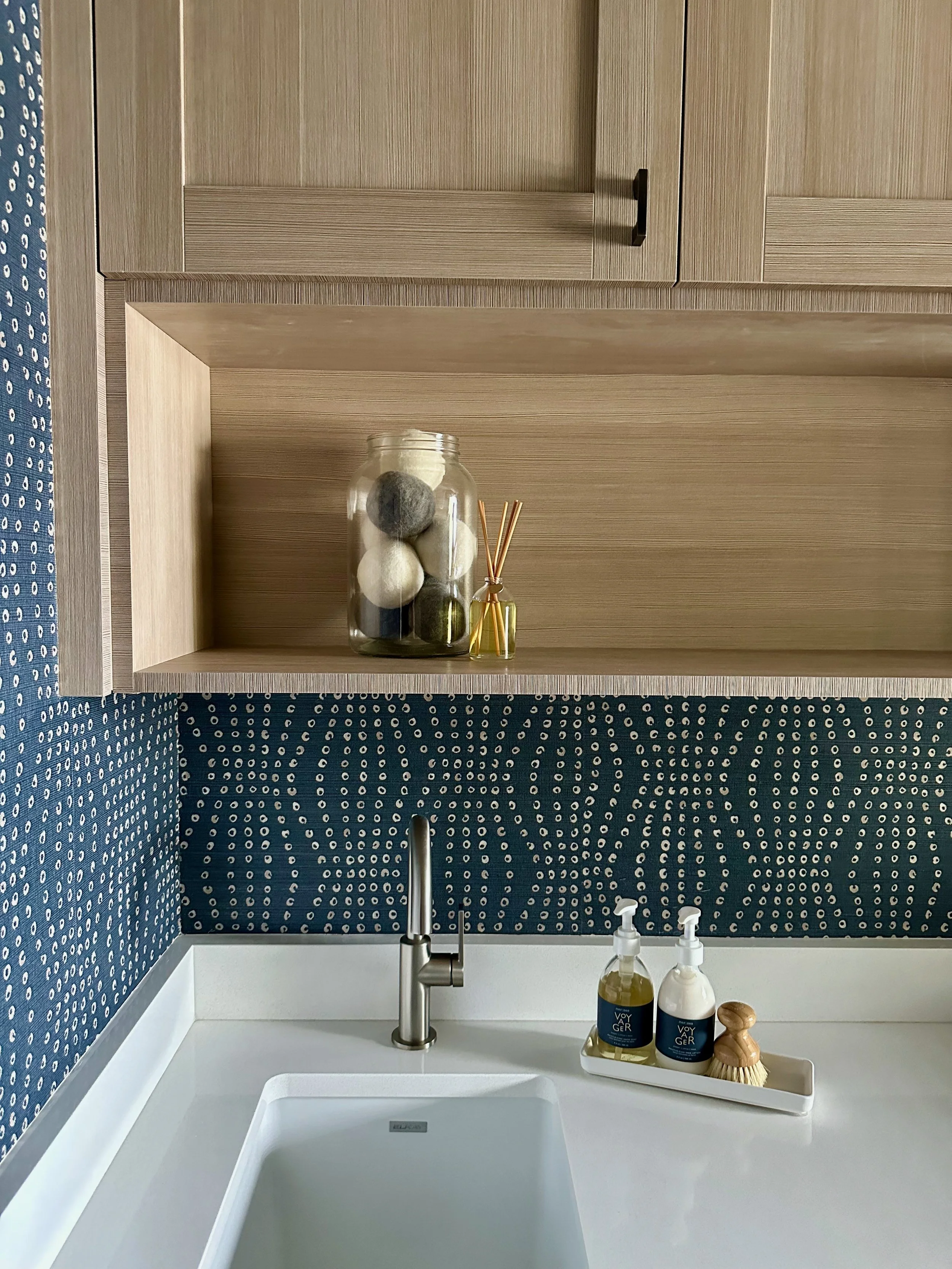 Modern kitchen sink area with a white countertop, blue patterned wallpaper, and a light wood cabinet. Features a silver faucet, glass jar with wool balls and reed diffuser, and soap dispensers on a tray.