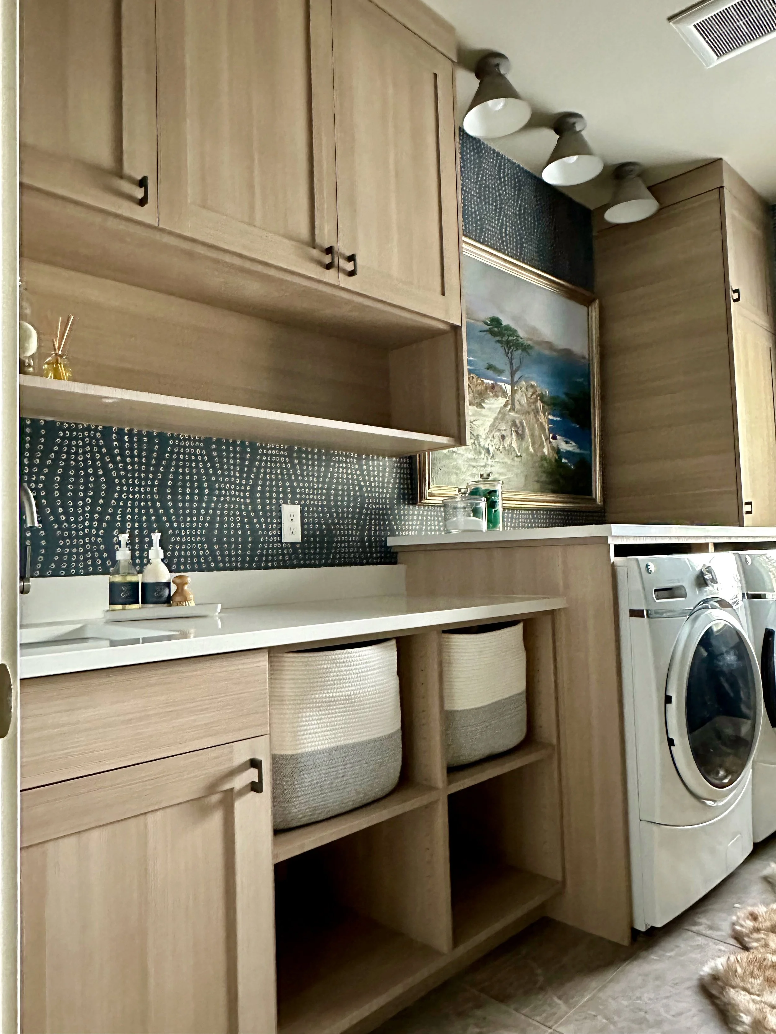 Laundry room with light wood cabinetry, a front-load washer and dryer, baskets for storage, a white countertop, wall art, and decorative lighting.