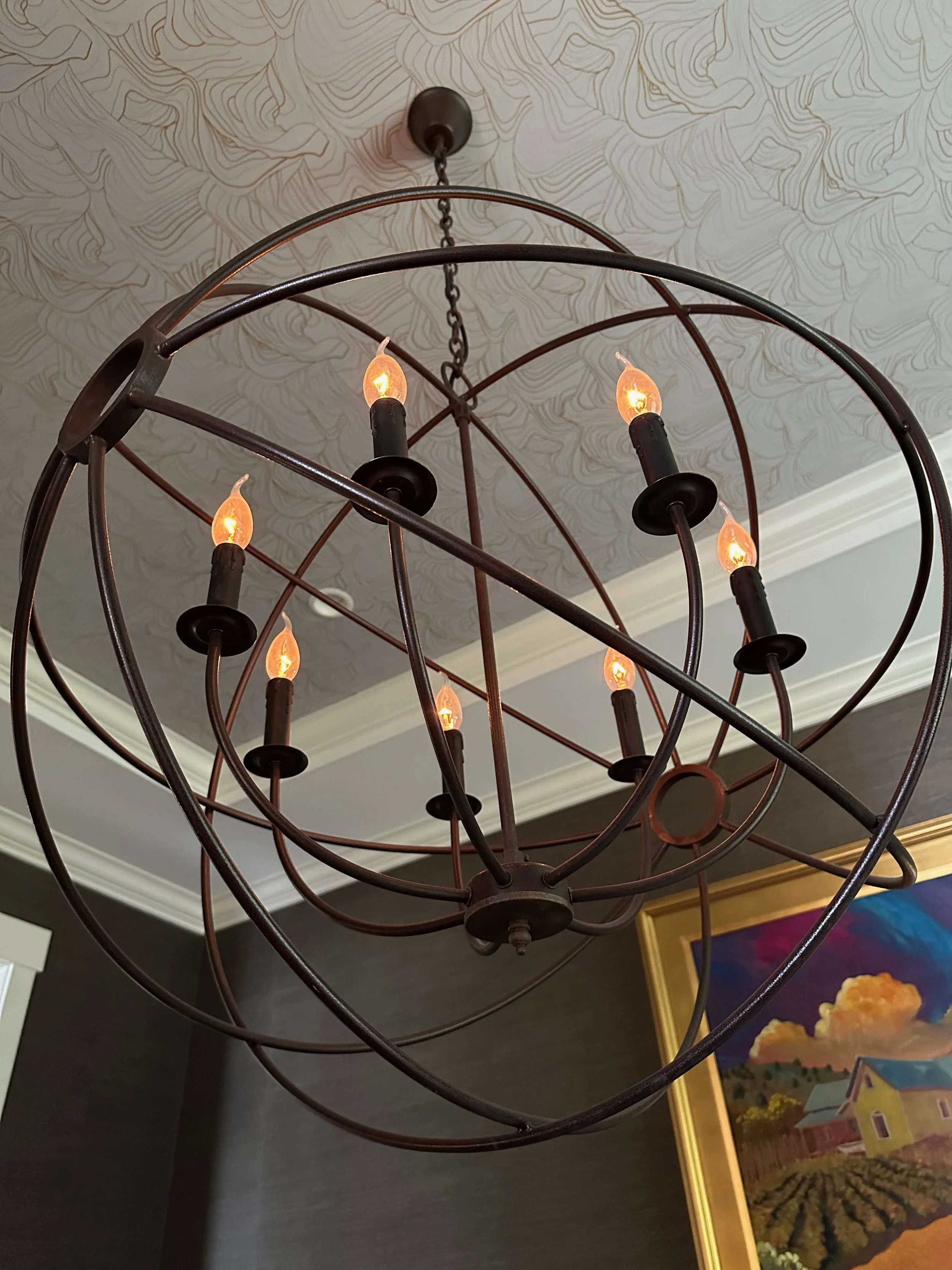 Orb-shaped metal chandelier with exposed Edison bulbs hanging from the ceiling, featuring a globe-like design. It is in a room with textured wallpaper and a colorful painting on the wall.