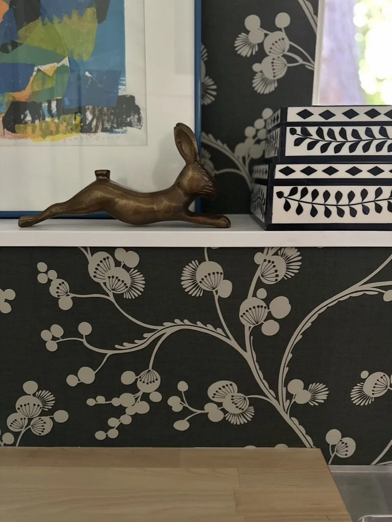 Decorative shelf with a bronze rabbit sculpture, abstract framed artwork, black and white patterned storage boxes, and floral wallpaper.