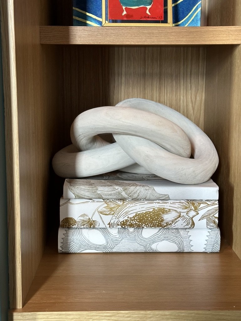 Wooden shelf with a decorative knot sculpture on top of a stack of books wrapped in white and gold patterned paper.