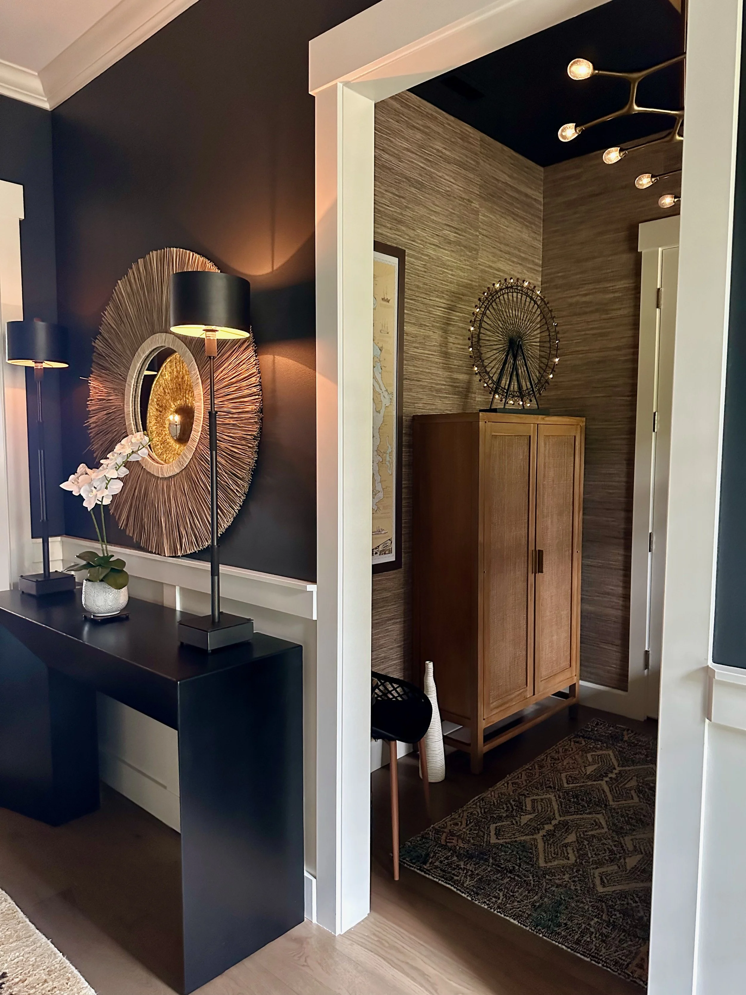 Stylish modern interior with black accent wall, decorative mirror, tall lamps, and black console table with orchids. Adjacent room features wooden cabinet, abstract art piece, and unique lighting fixture on ceiling.