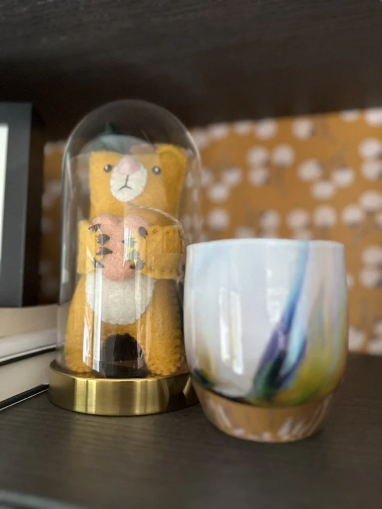 Stuffed bear ornament under glass cloche next to a colorful ceramic cup on a shelf.