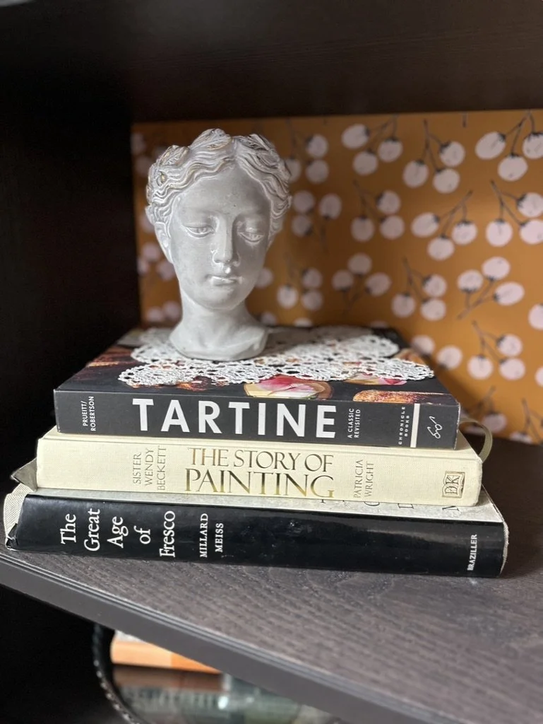 Stack of books with a small bust sculpture on top, set against floral wallpaper background.