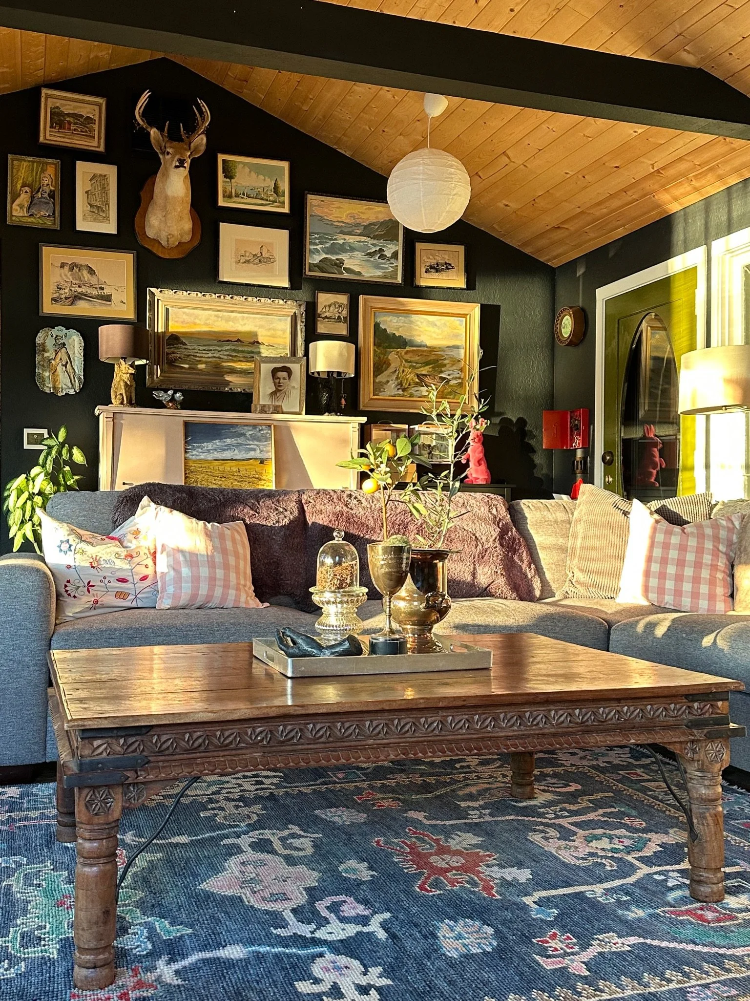 Cozy living room with a gray sofa decorated with pink and white pillows, large wooden coffee table, rich patterned rug, and a gallery wall featuring various framed artworks and a mounted deer head. The room has a vaulted wooden ceiling, warm lighting