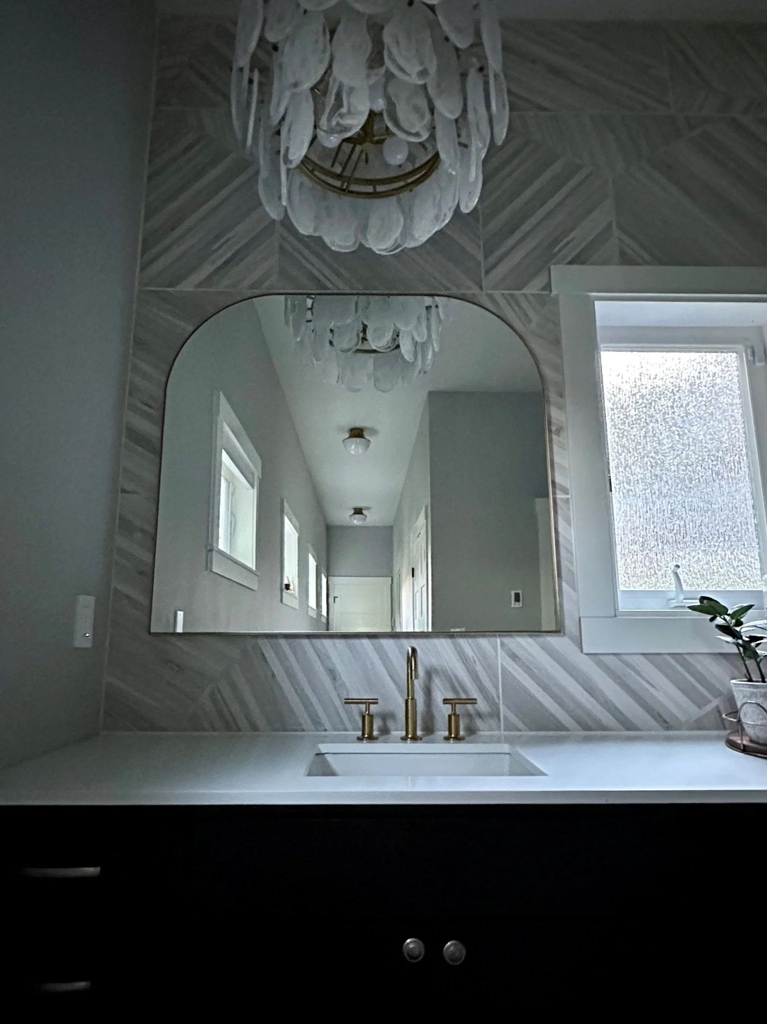 Modern bathroom vanity with a white countertop, brass faucet fixtures, and a tiled backsplash. A large mirror reflects ceiling lights, and there is a small potted plant on the right side of the counter.