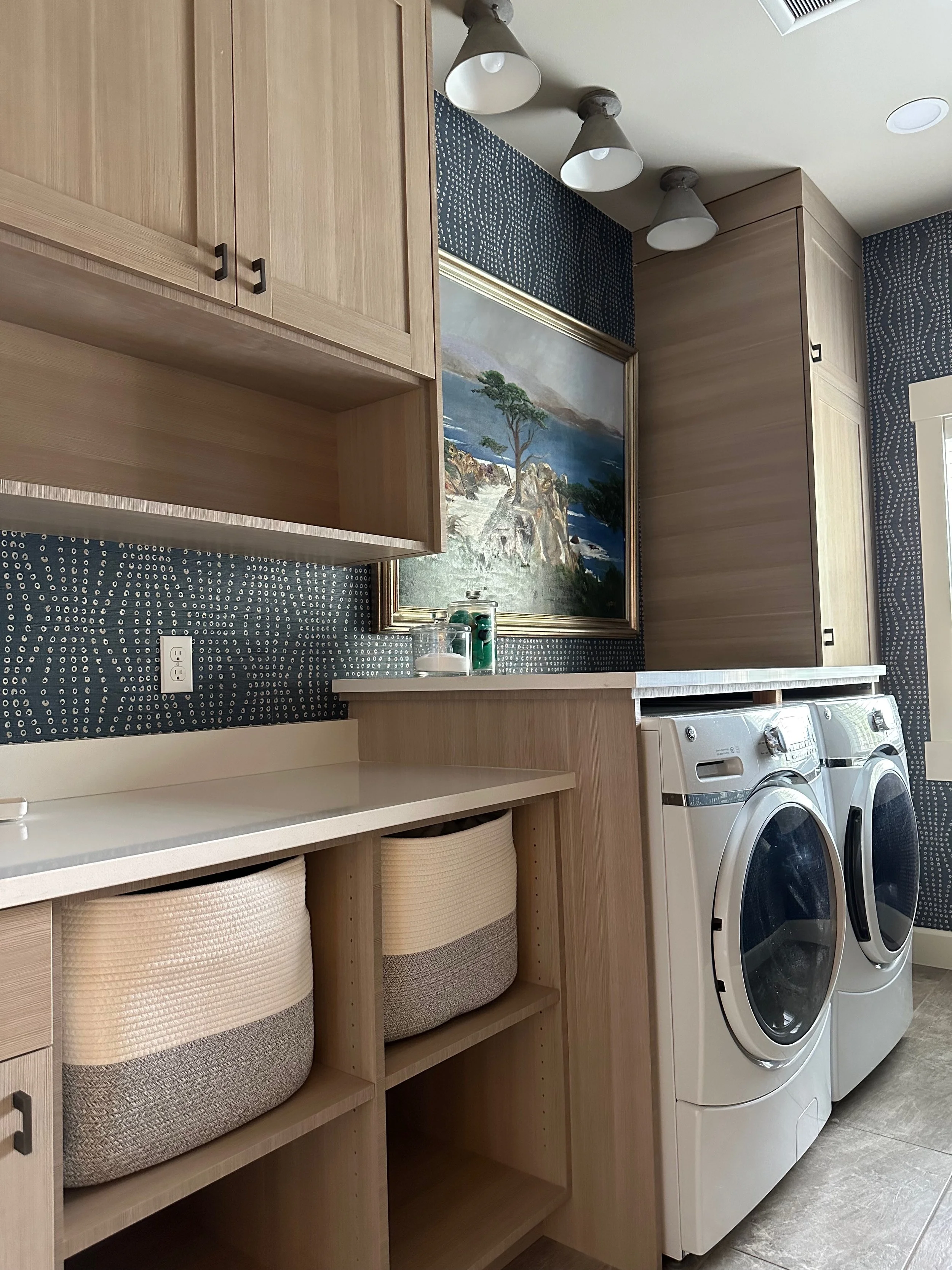 A modern laundry room with wood cabinets, front-loading washer and dryer, countertop space, decorative wallpaper, and a framed landscape painting.