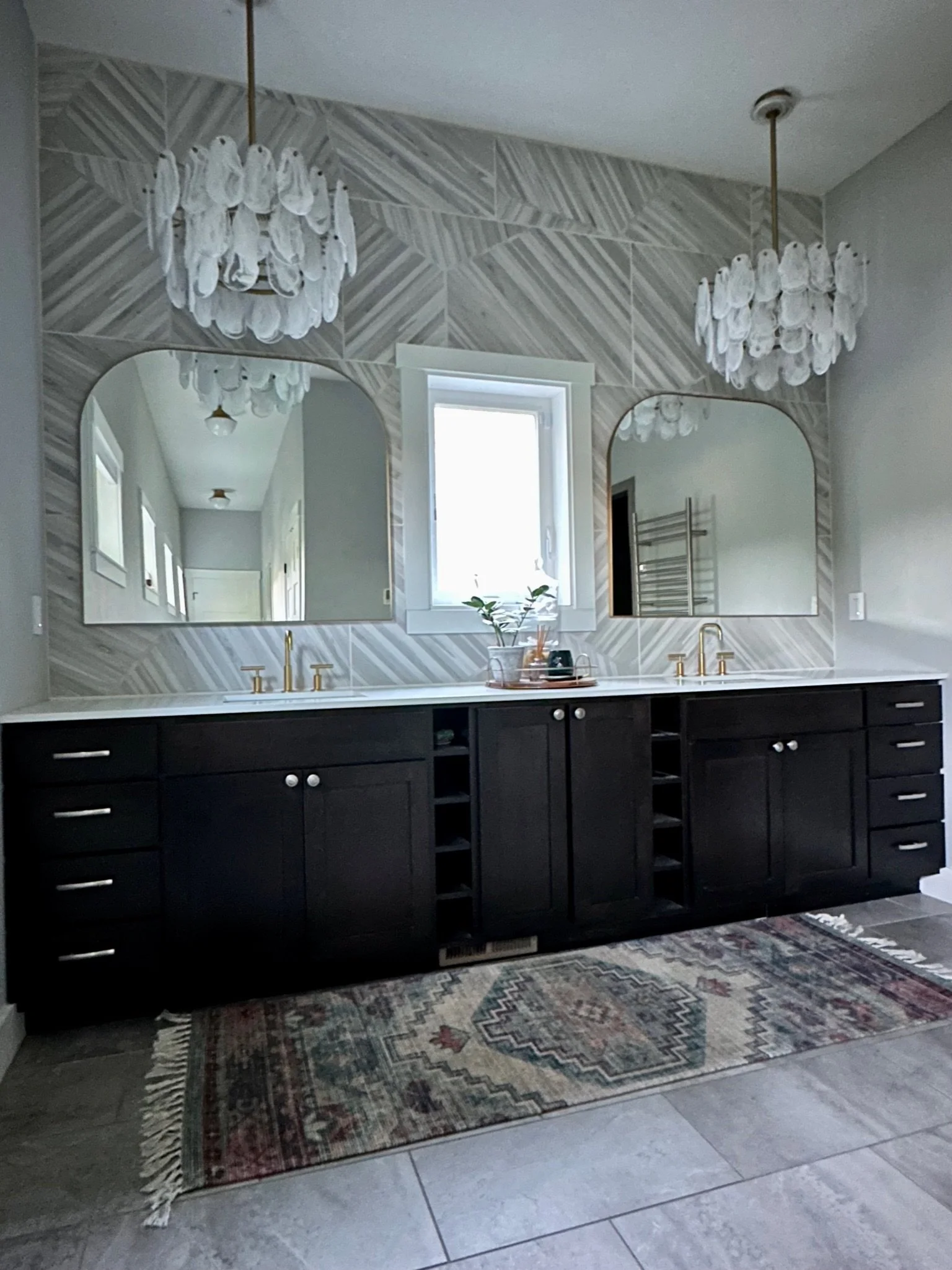 Modern bathroom with dual sinks, dark wood vanity, gold faucets, arched mirrors, geometric wall tiles, hanging chandeliers, small window, and a decorative rug on tiled floor.
