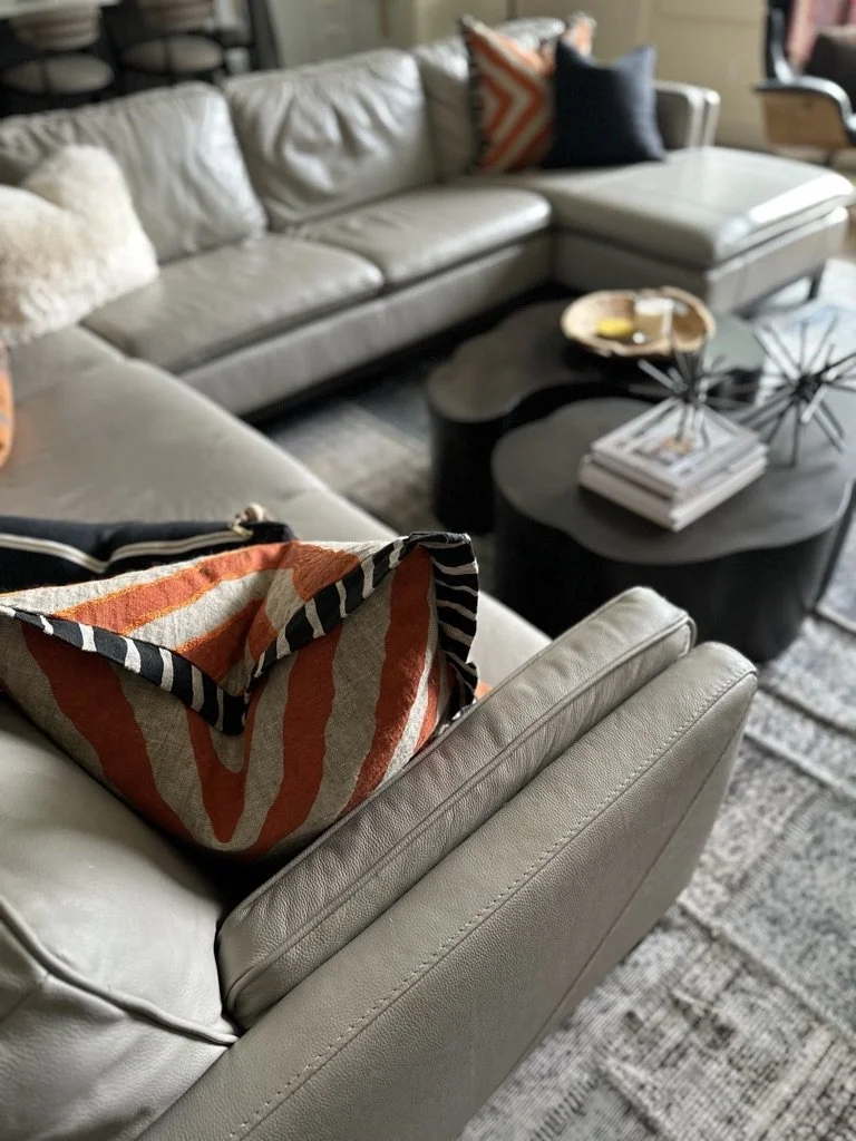 Modern living room with gray sectional sofa, colorful throw pillows, nesting coffee tables, decorative sculpture, and stacked books.