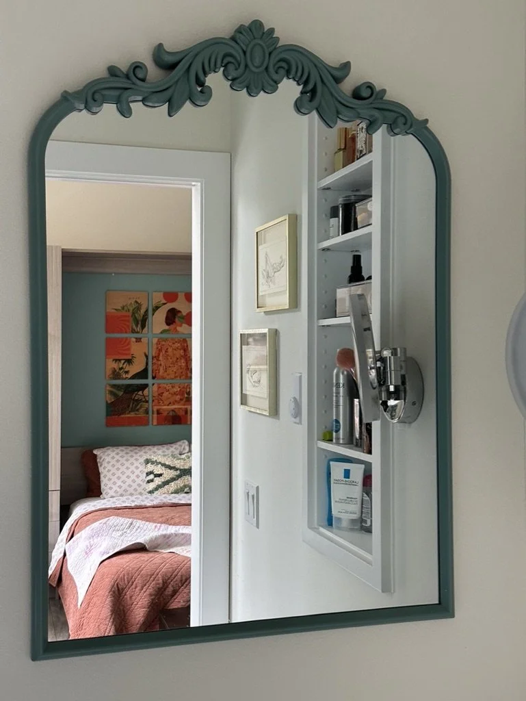 Ornate mirror reflecting a bedroom, with shelves holding personal items beside it.