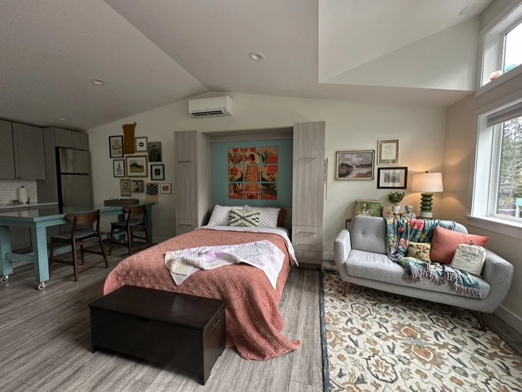Interior of a cozy studio apartment featuring a bed with colorful pillows and blanket, a small gray couch with decorative pillows and a throw, a rug with floral patterns, and some wall art. In the background, there's a kitchen area with a counter and
