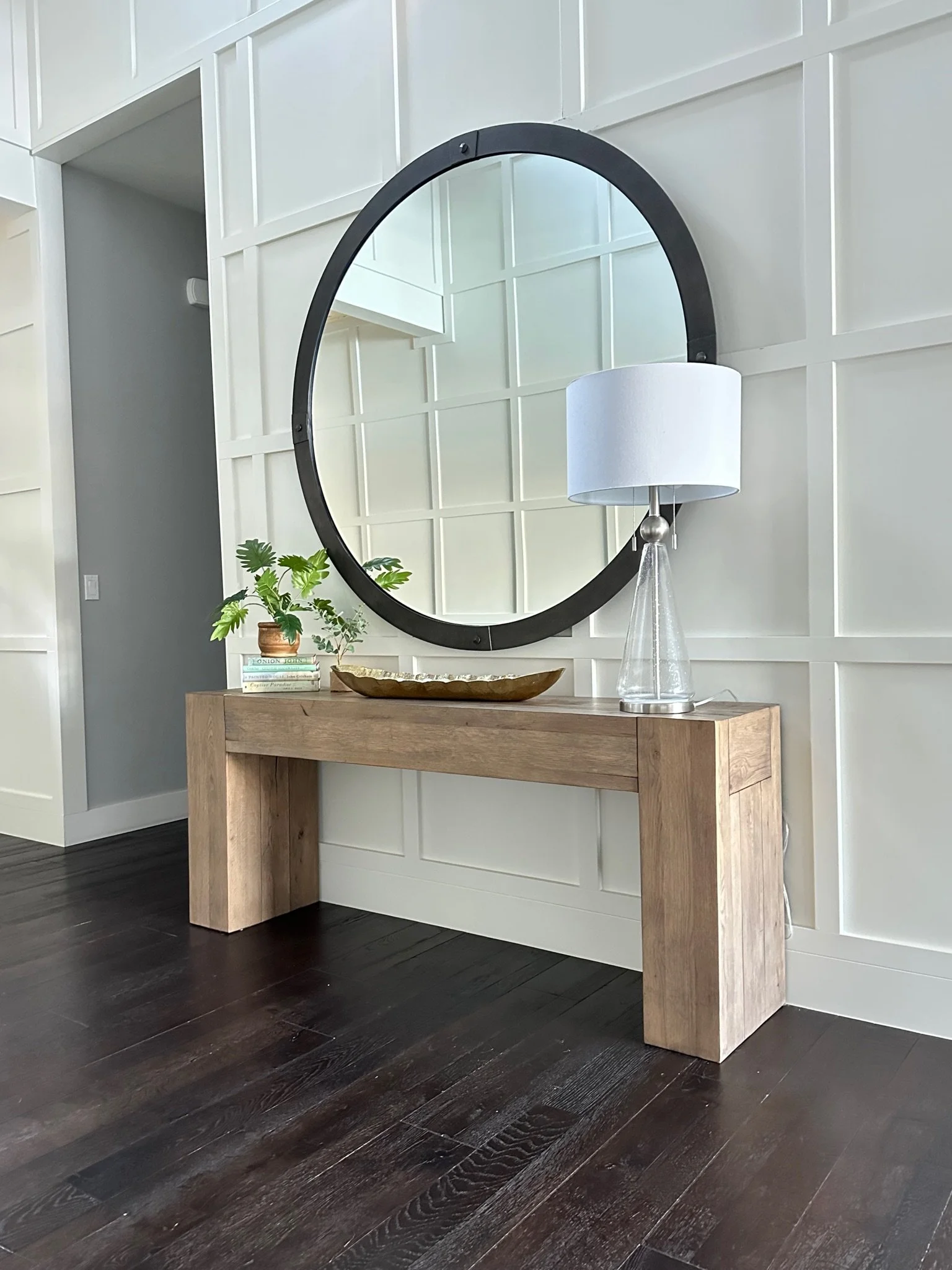Modern entryway with a wooden console table, large round mirror, table lamp, decorative bowl, and potted plant on dark wood flooring.