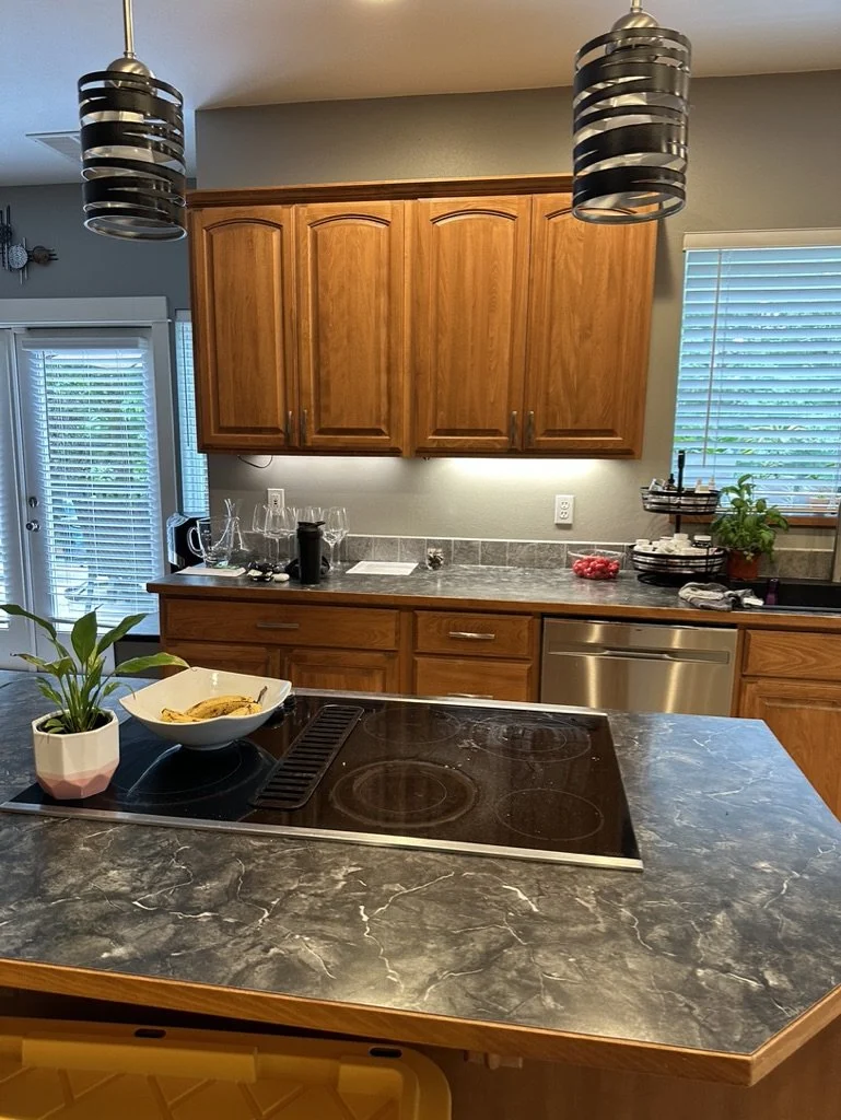 Modern kitchen with wooden cabinets, marble countertops, an electric stovetop, pendant lighting, a bowl of bananas, a potted plant, and a stainless steel dishwasher.
