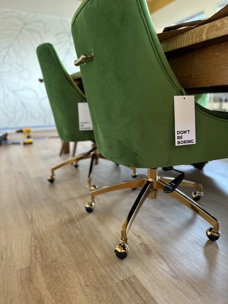 Green swivel chairs with gold legs on a wooden floor, each with a tag reading "Don't be boring."