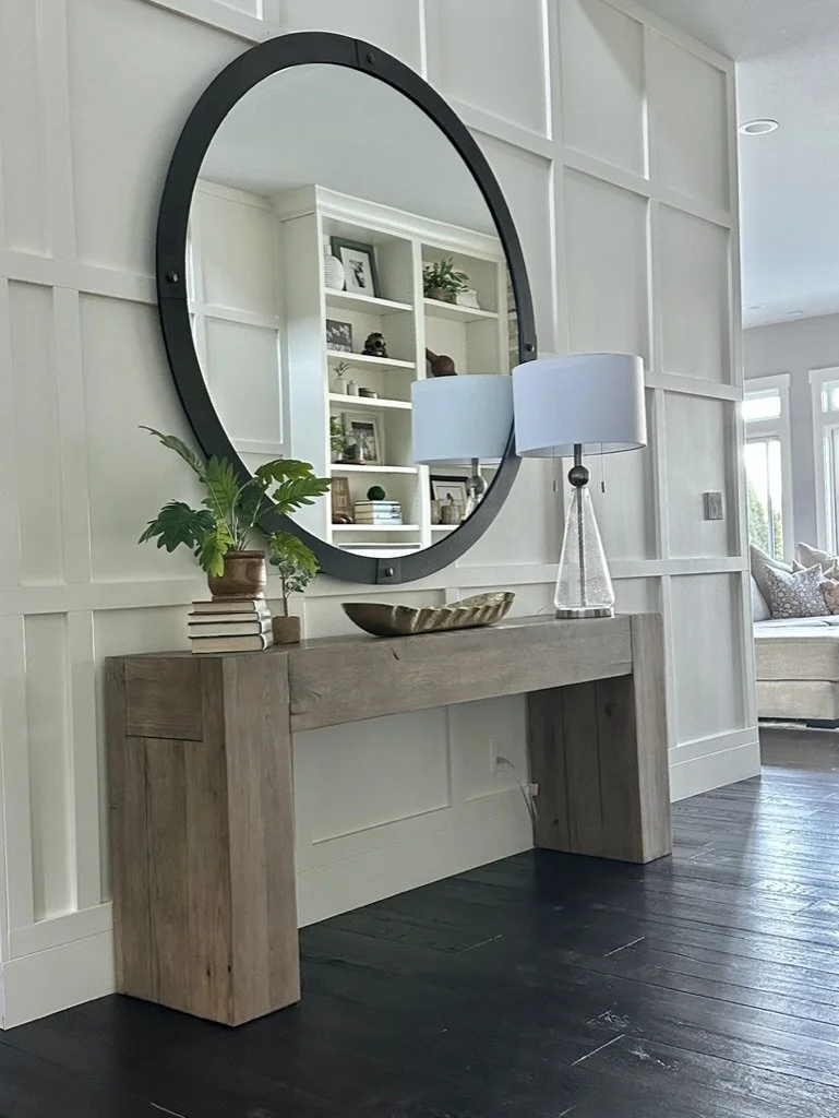A modern foyer features a large round mirror mounted on a paneled wall. Below it is a rustic wooden console table with a plant, a stack of books, and a decorative bowl. A lamp with a round shade is placed on the table, and shelves with decor are visi