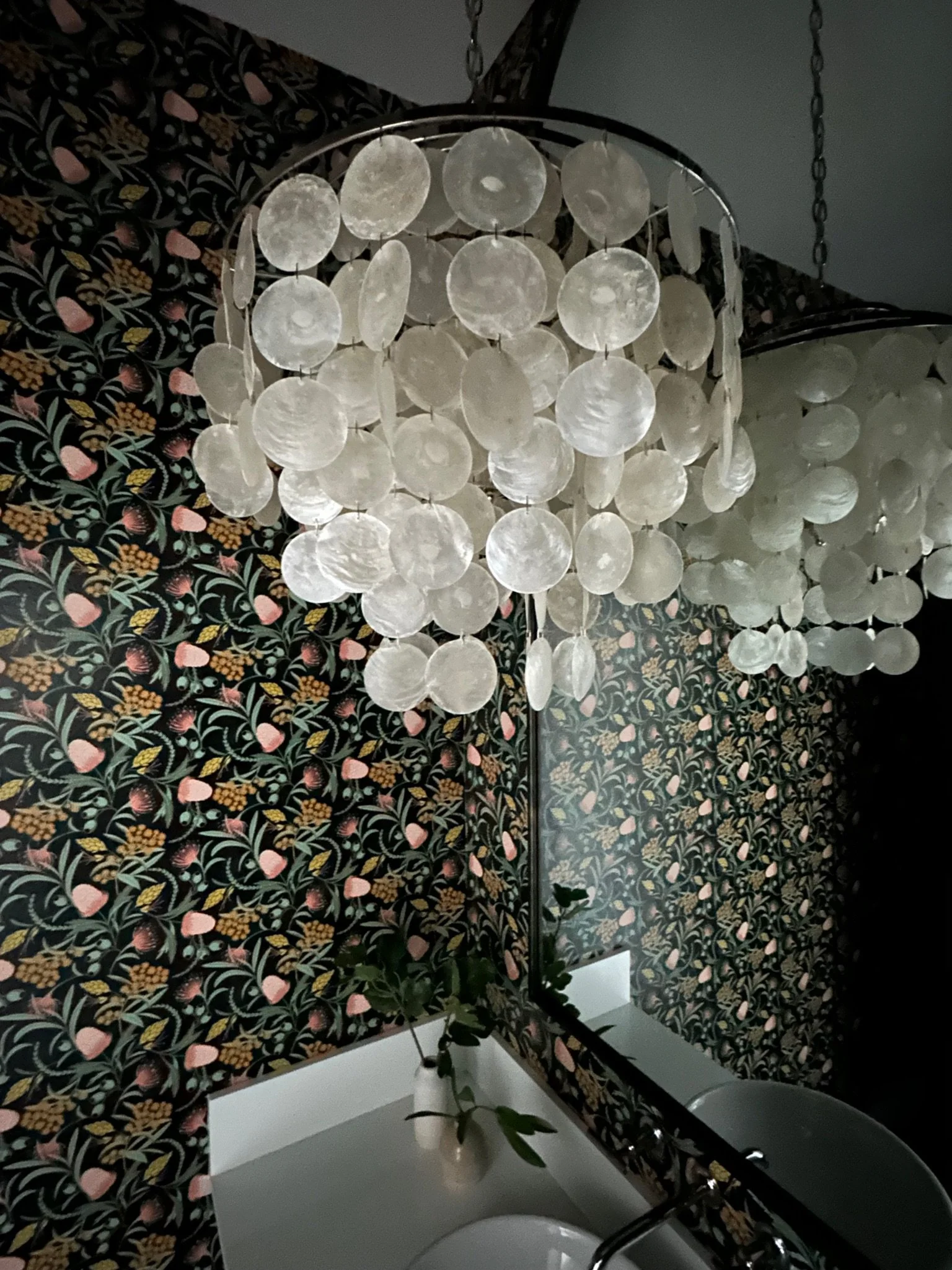 A decorative chandelier with capiz shells hangs in a room with vibrant floral wallpaper. Below the chandelier is a countertop with a white vase containing green leaves, reflected in a large mirror.