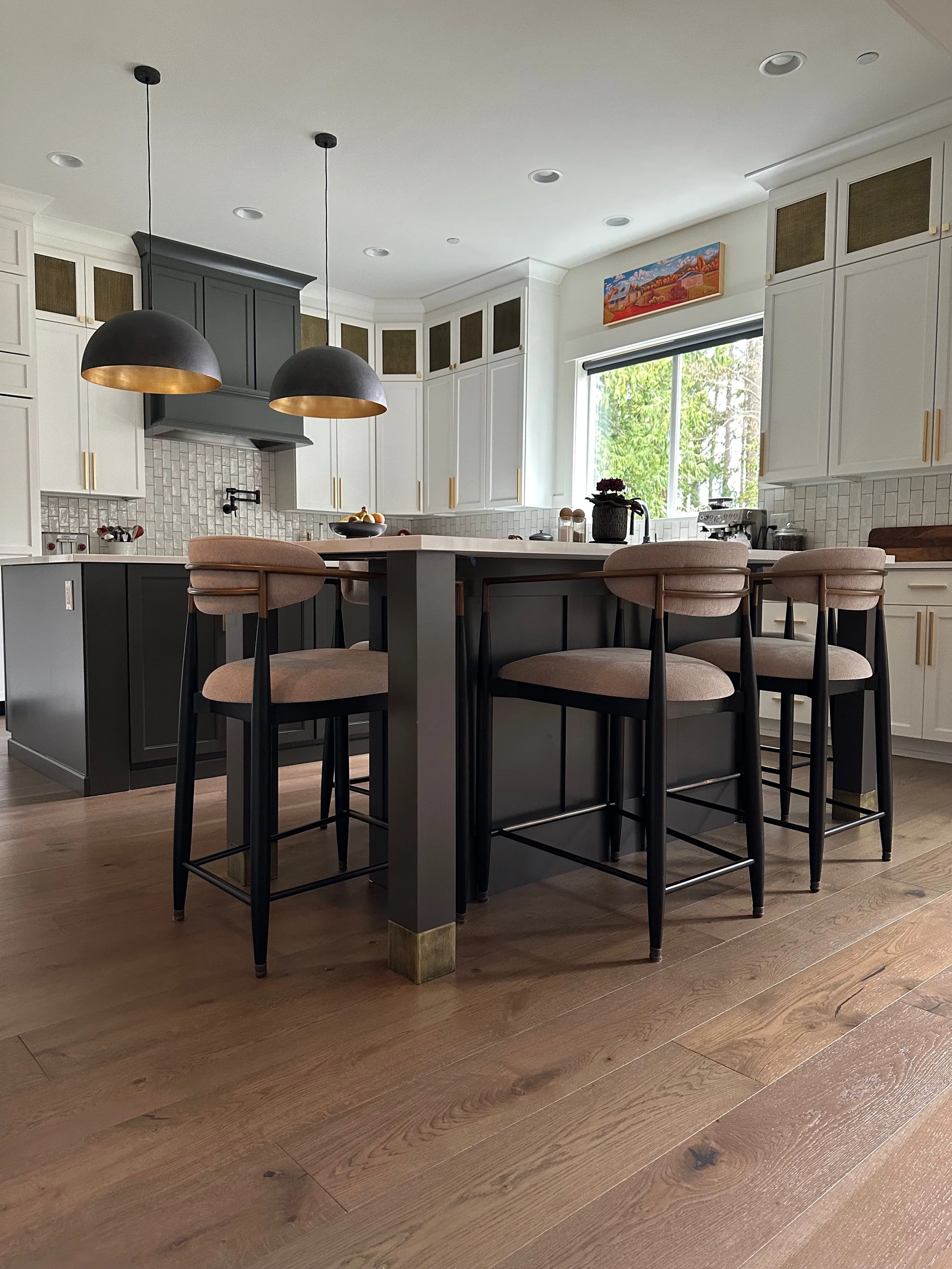 Modern kitchen with a large island featuring dark cabinetry, light countertops, and pendant lighting. The room has hardwood flooring and white cabinets with brass handles. There are four upholstered bar stools, and a window with a view of trees. A co