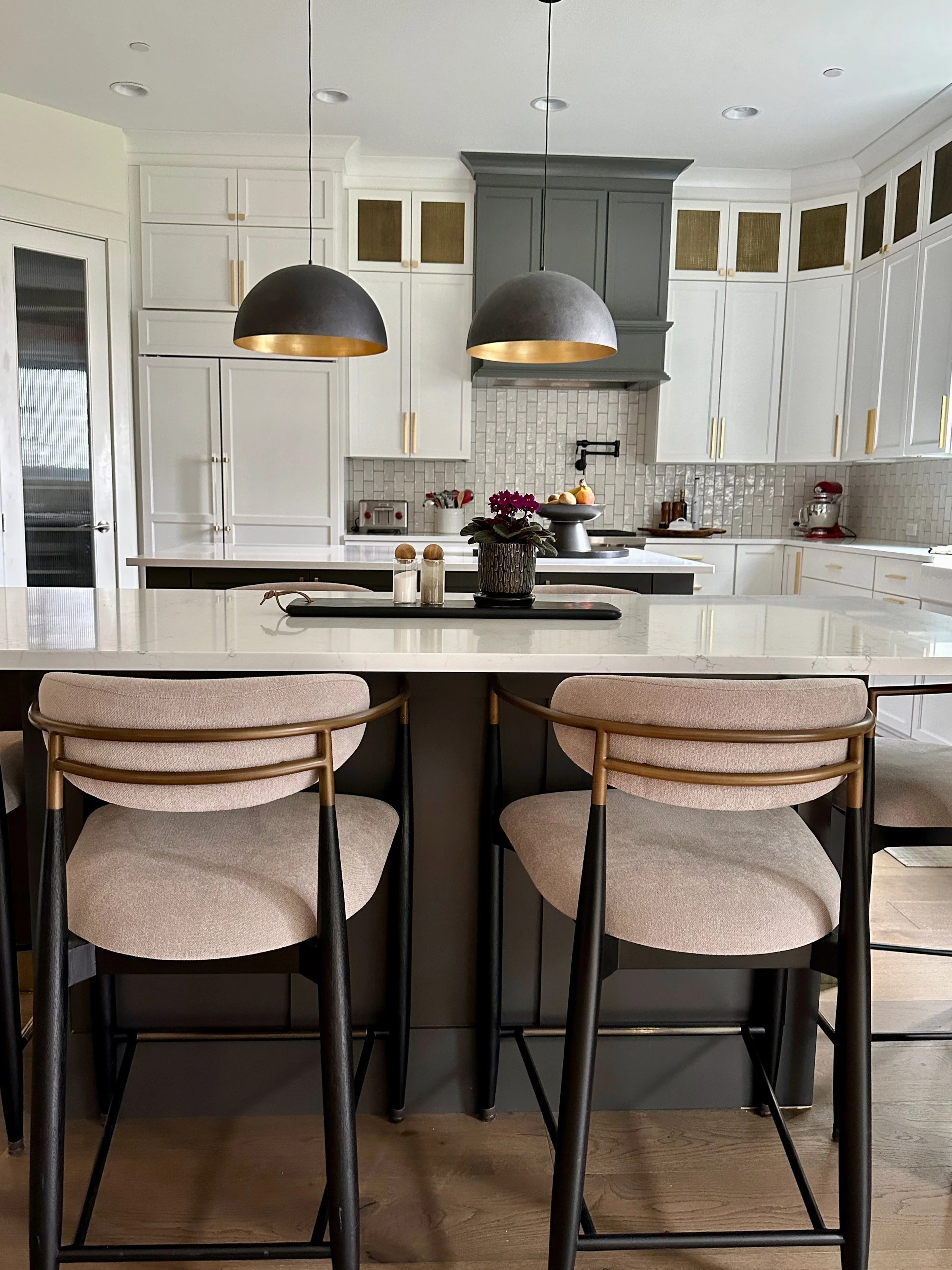 Modern kitchen interior with white cabinets, central island, and two pendant lights. Features bar stools, a plant on the counter, and a tiled backsplash.