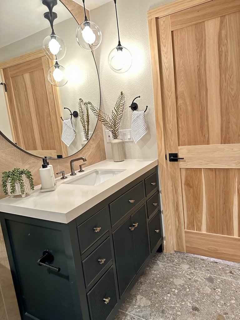 Bathroom interior with a wooden door, dark vanity with a white sink, round mirror, pendant lights, potted plant, and hand towel.