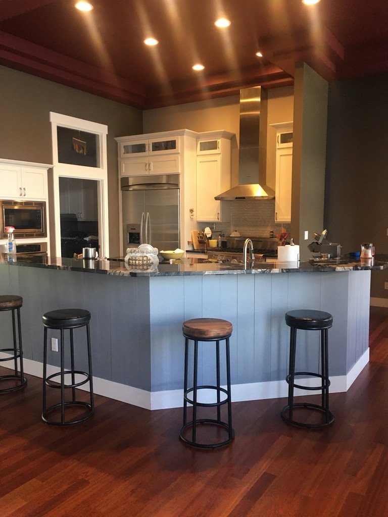 Modern kitchen with white cabinets, stainless steel appliances, a large range hood, and a granite-topped island with bar stools on a wooden floor.