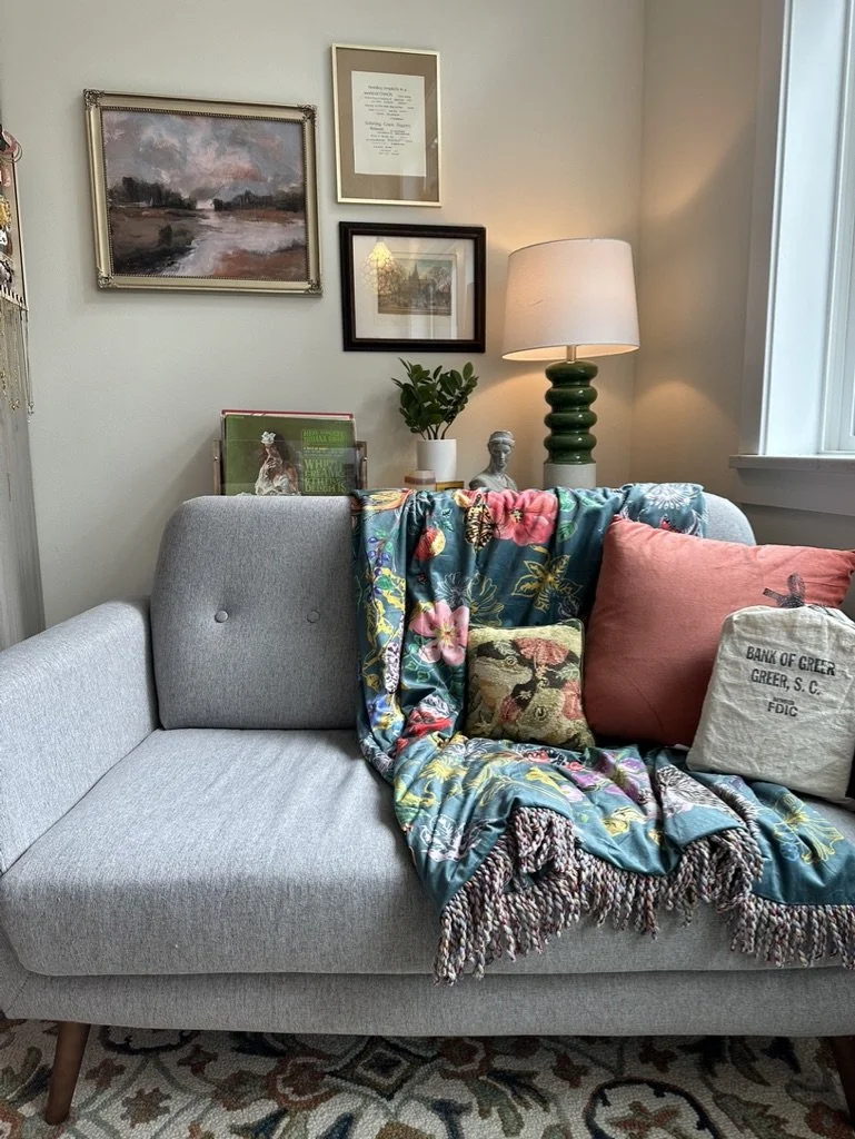 Cozy living room corner with a gray sofa, colorful floral blanket, decorative pillows, green table lamp, and framed artwork on the walls.