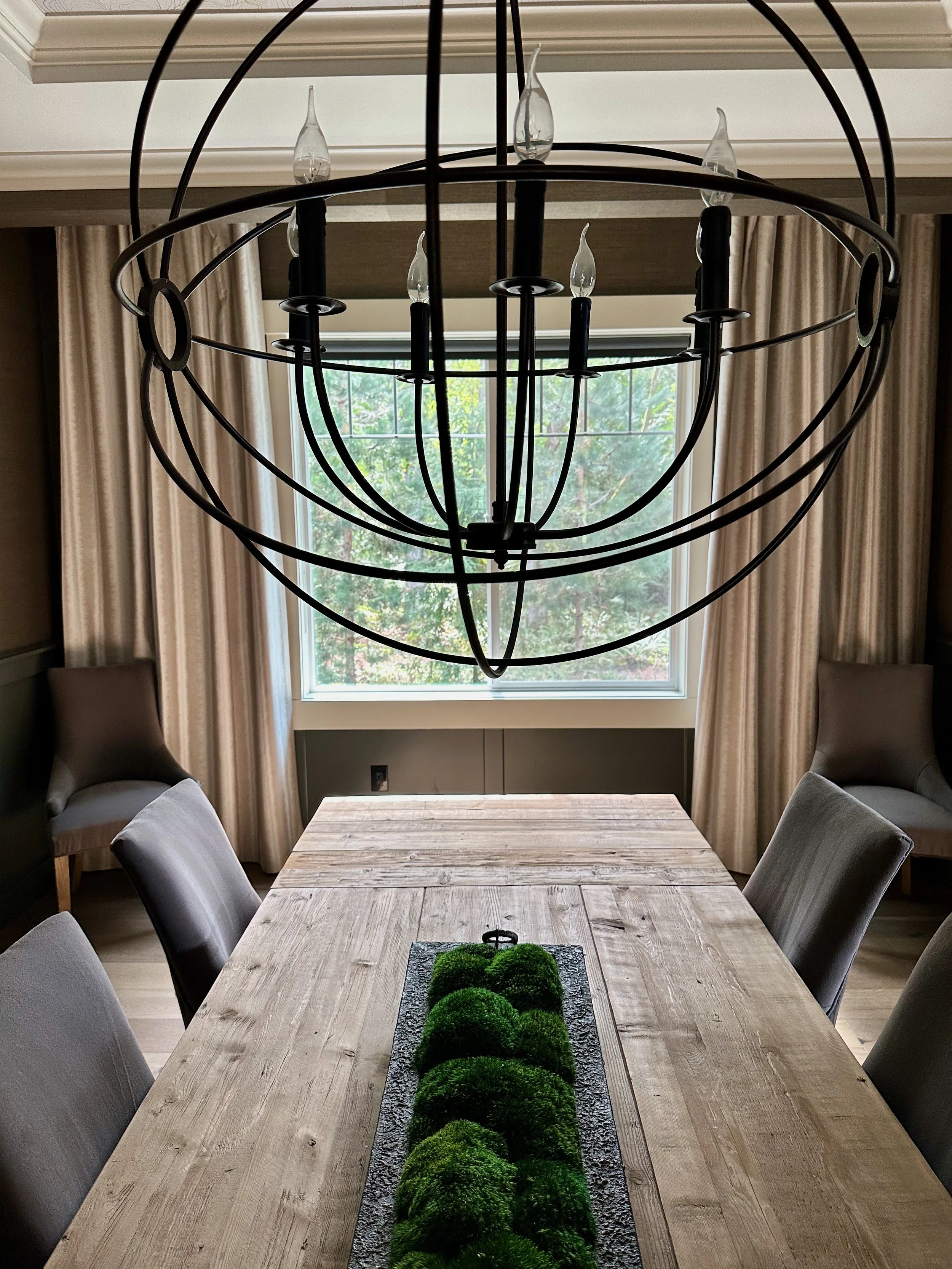 Dining room with wooden table, modern chandelier, and green moss centerpiece. Four gray upholstered chairs around the table, with curtains and a window in the background.