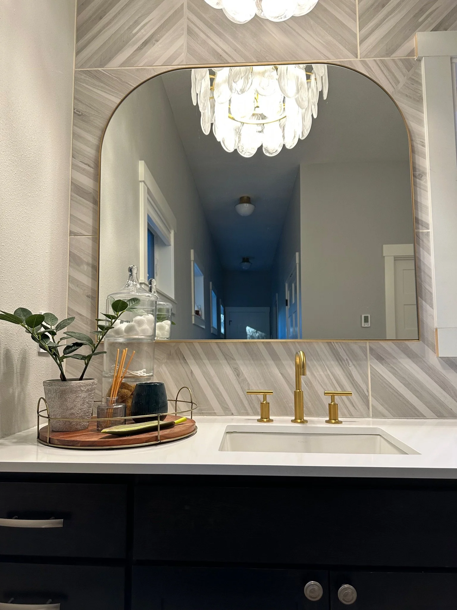 Modern bathroom vanity with a gold faucet, white countertop, arched mirror, gray textured tile backsplash, decorative wall sconce, plant in a pot, glass jars with cotton balls, and a fragrance diffuser on a round tray.