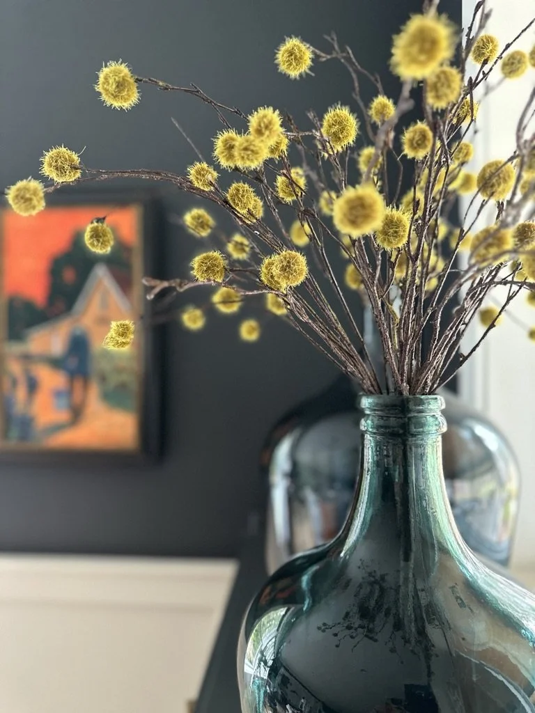 Decorative arrangement with yellow pom-pom flowers in a dark green glass vase, against a dark wall with an abstract painting.