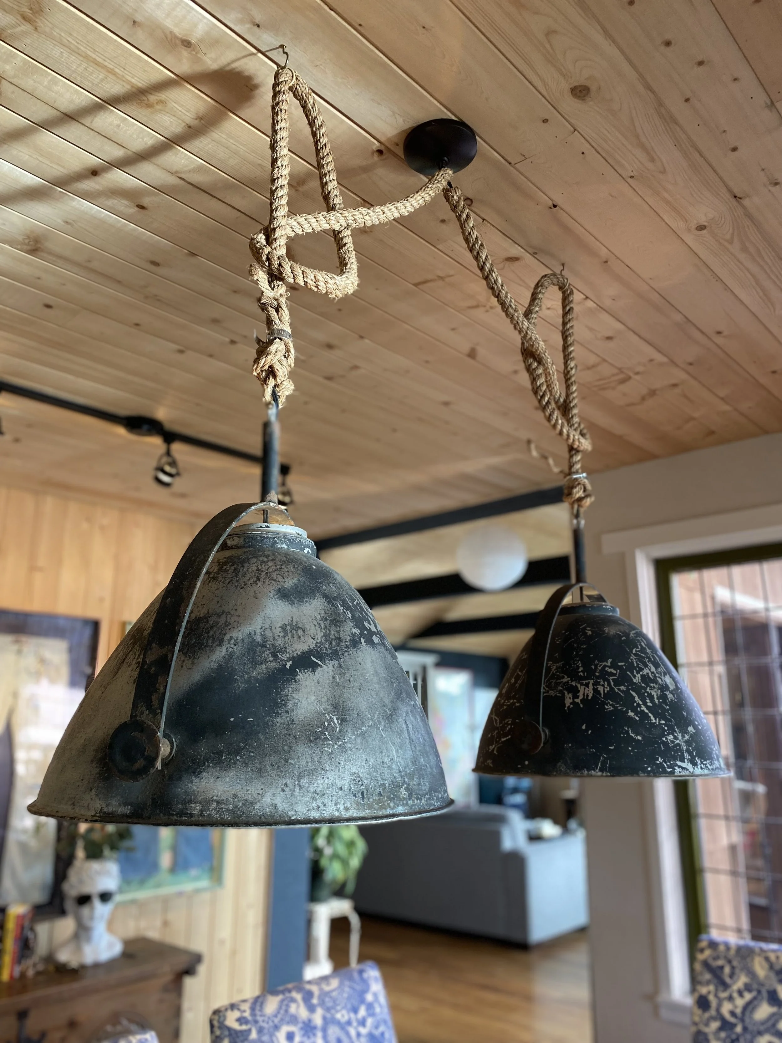 Rustic pendant lights with rope hanging from a wooden ceiling in a cozy room, featuring artwork and a bust on a table.