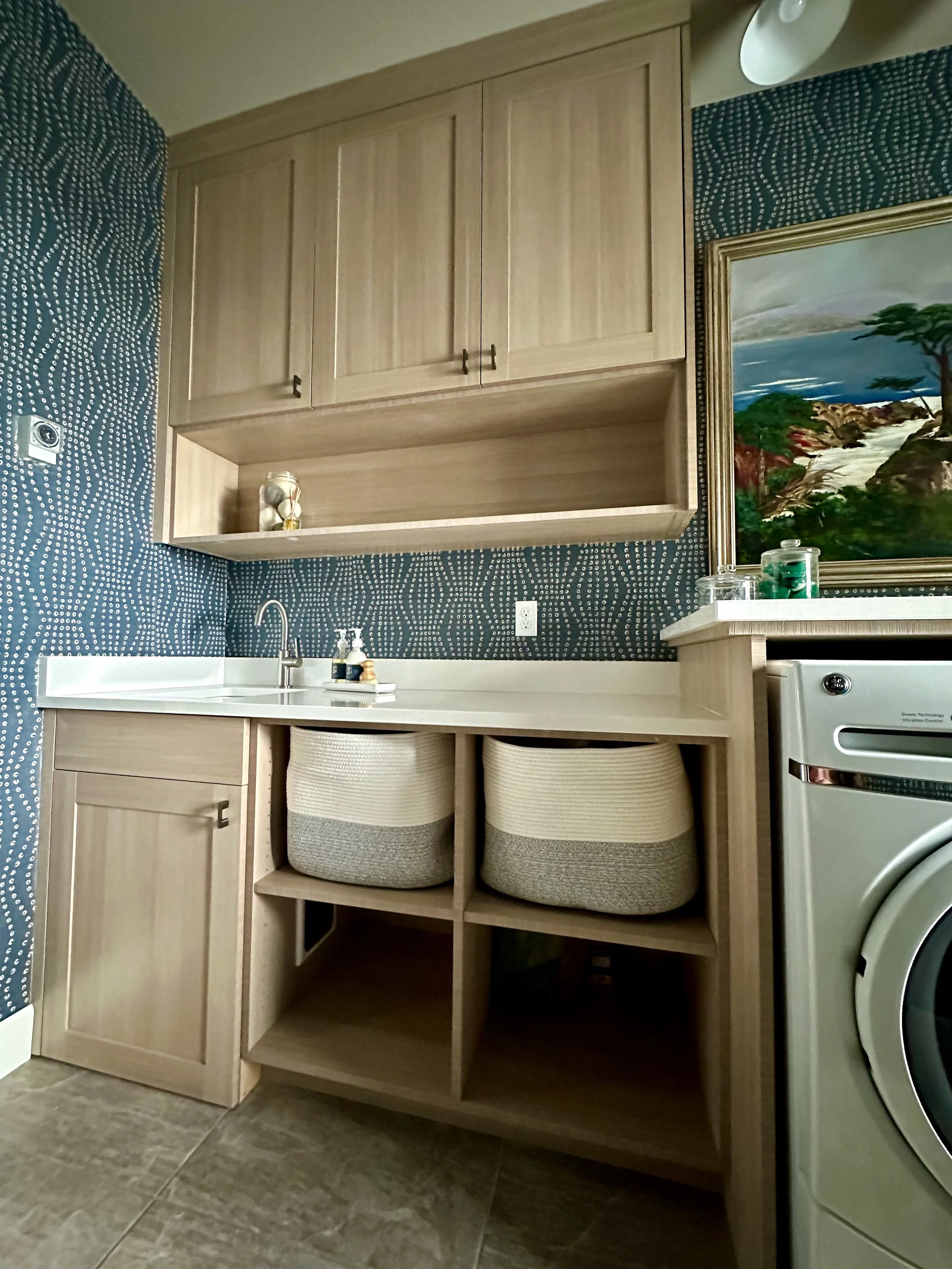 Laundry room with beige cabinets, a blue patterned wall, a countertop sink, storage baskets, and a painting. A washing machine is partially visible on the right.