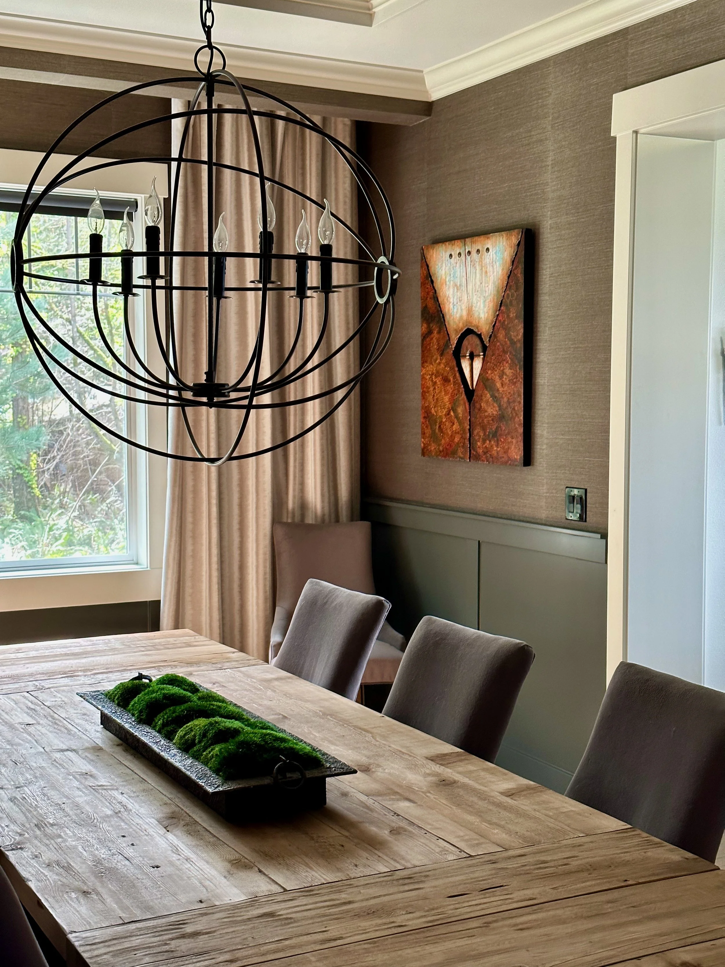 Dining room with modern chandelier, wooden table, cushioned chairs, tray with green moss centerpiece, abstract wall art, and large window.