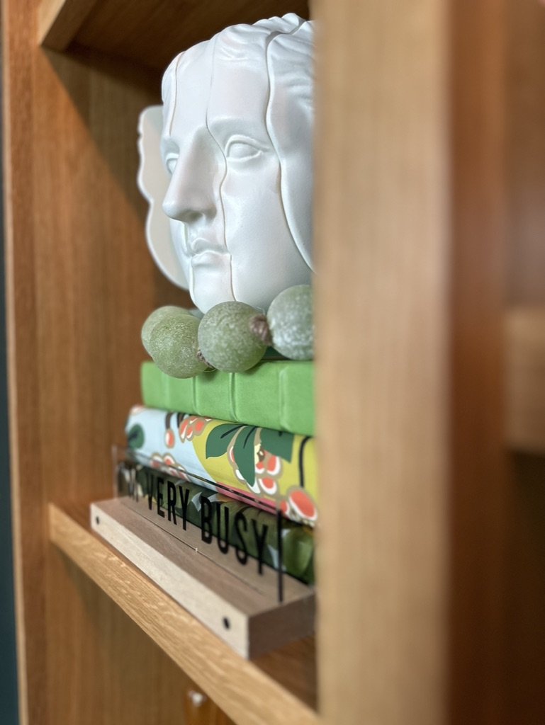 Close-up of a decorative white sculpted face on a shelf, alongside stacked books with colorful covers, and a small sign saying "VERY BUSY."